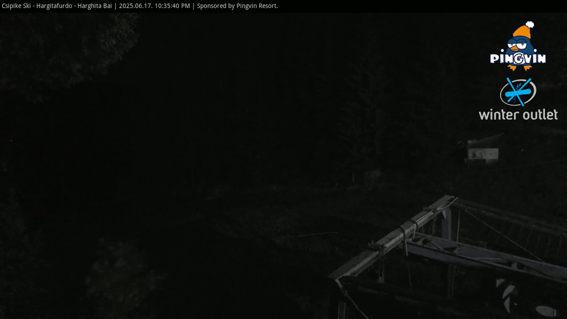 A nighttime, dark view shows a portion of a structure, possibly a ski lift, partially visible against a very dark background of trees.