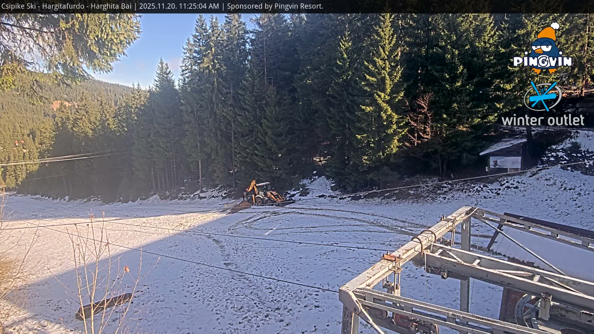 A sunny winter day reveals a snowy mountain landscape with a dense evergreen forest, a yellow backhoe working on a partially snow-covered slope, and parts of a ski lift mechanism in the foreground.