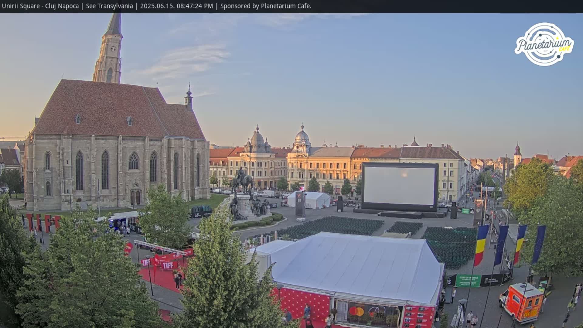 A large city square under a clear, evening sky features a church, several buildings, a large screen, and numerous chairs set up for an outdoor event.