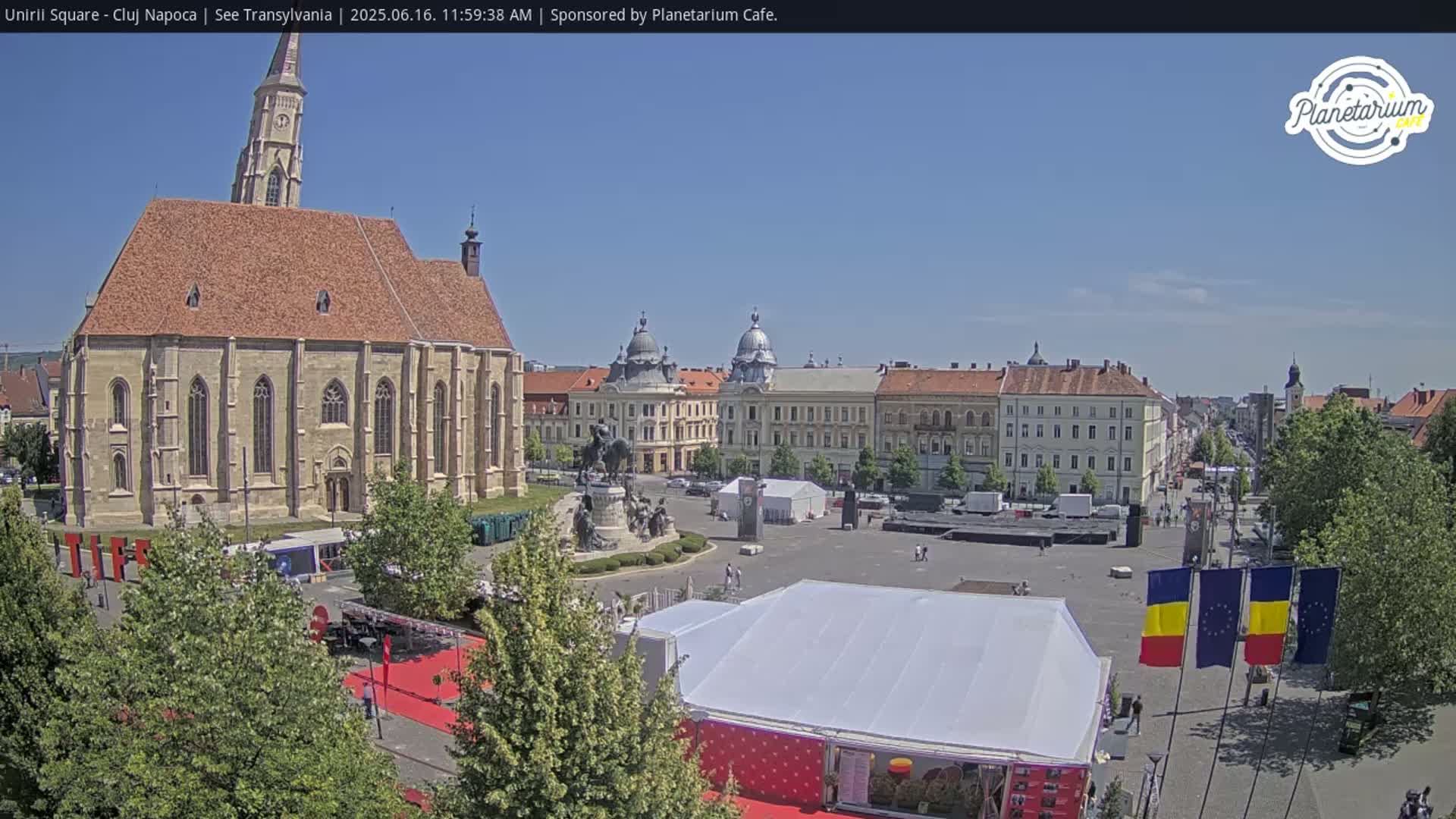 A large town square on a sunny day, featuring a prominent church, other buildings, a monument, and temporary event structures.