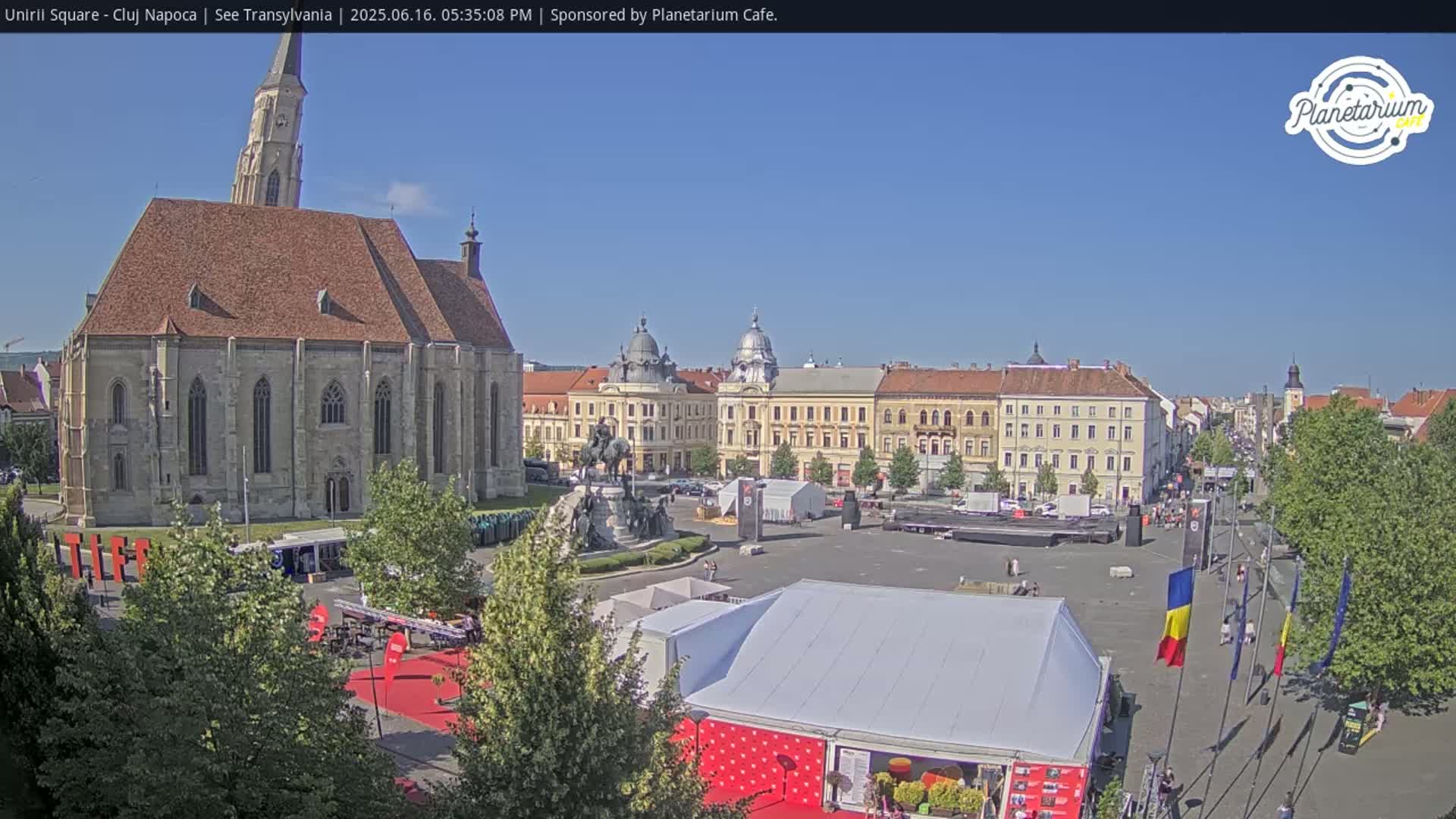 A large town square with a church, several buildings, a statue, and numerous tents under a clear, sunny sky.