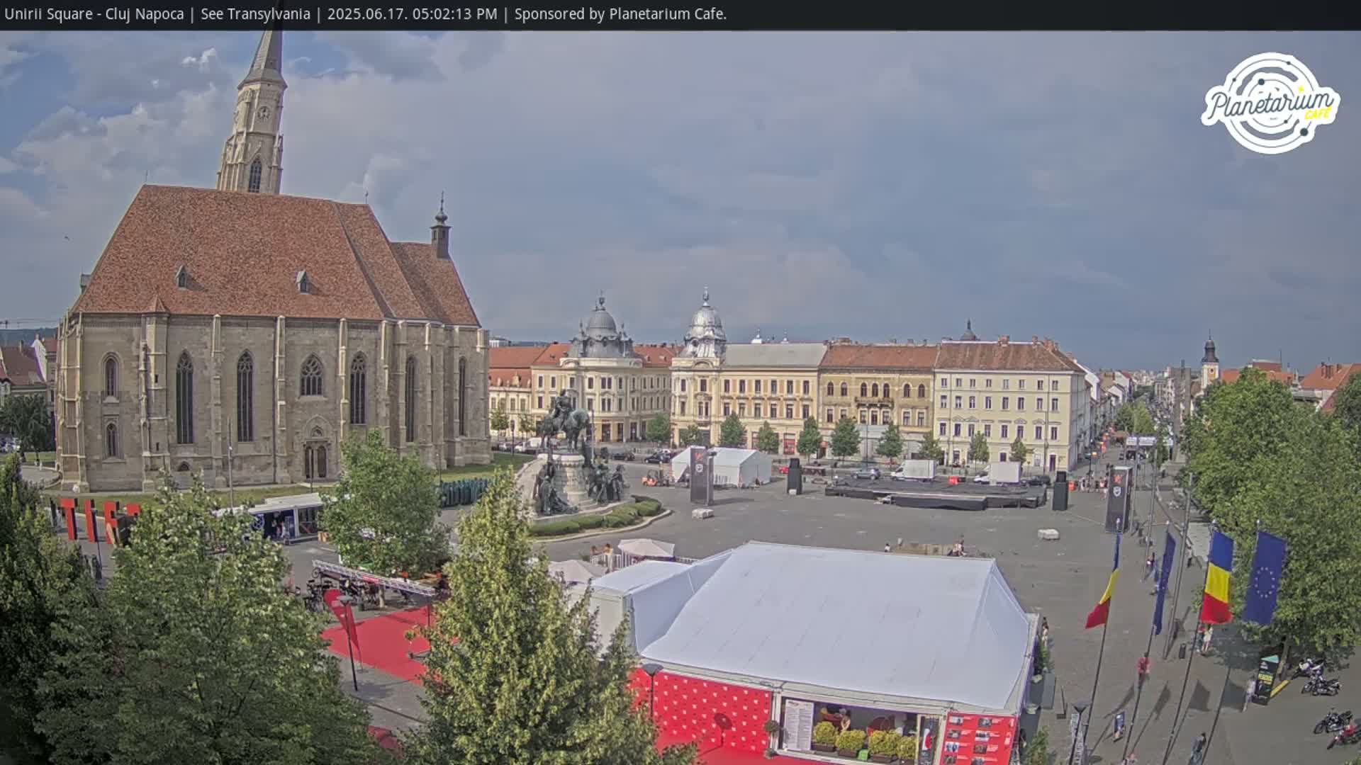 A large town square with a church, several buildings, a statue, and tents under partly cloudy skies.