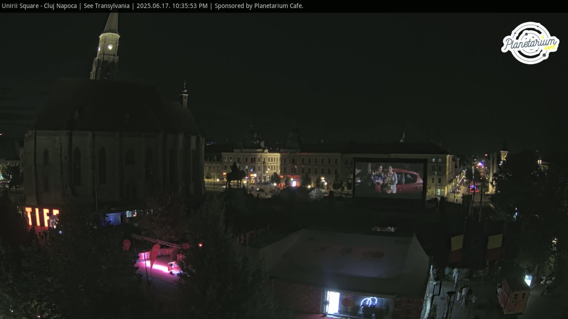 A nighttime aerial view of a city square showing a large building, several other structures, and an outdoor movie screen displaying a film.