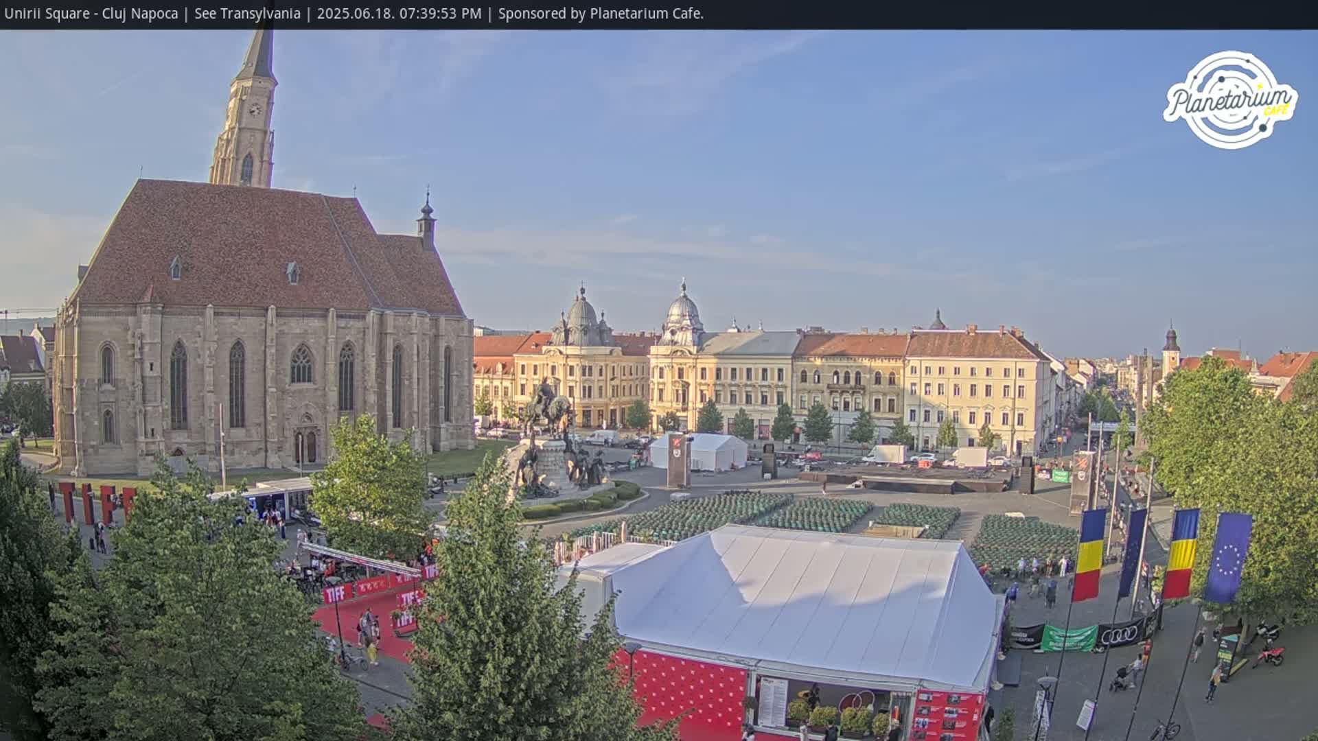 A large town square on a sunny day, featuring a large church, several buildings, a statue, and numerous tents set up in the square.