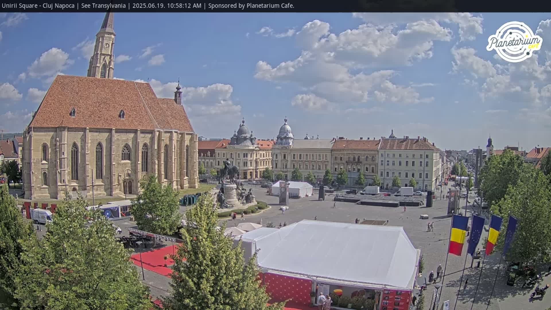 A large town square with a church, several buildings, a statue, and numerous tents under a partly cloudy sky.