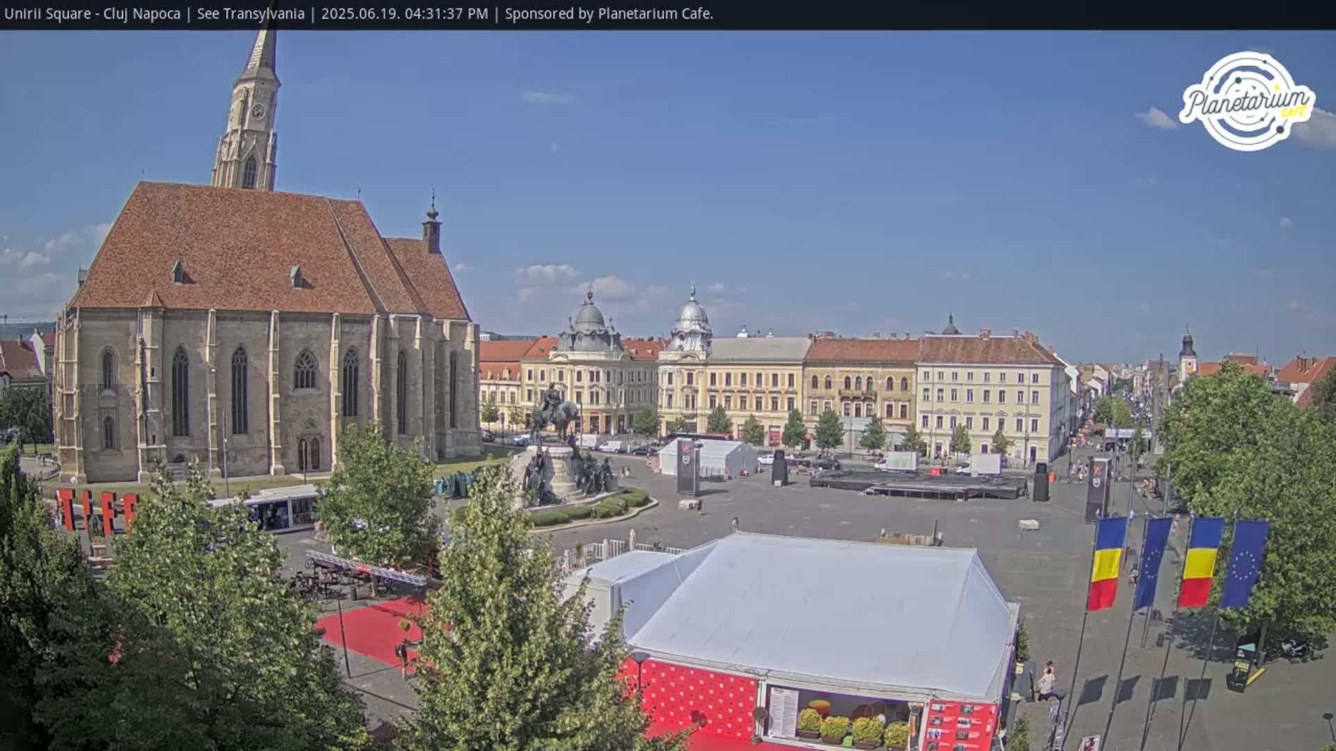 A large town square on a sunny day, featuring a prominent church, other buildings, a statue, and several tents set up in the plaza.