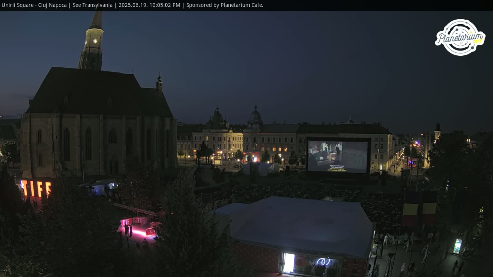 A nighttime outdoor movie screening in a town square, with a large church and other buildings visible in the background under a dark sky.