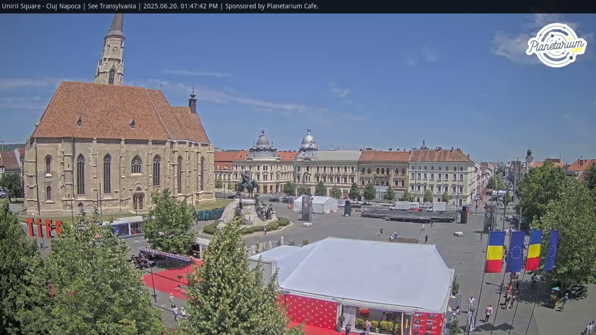A large town square on a sunny day features a large church, several other buildings, a statue, and several tents.