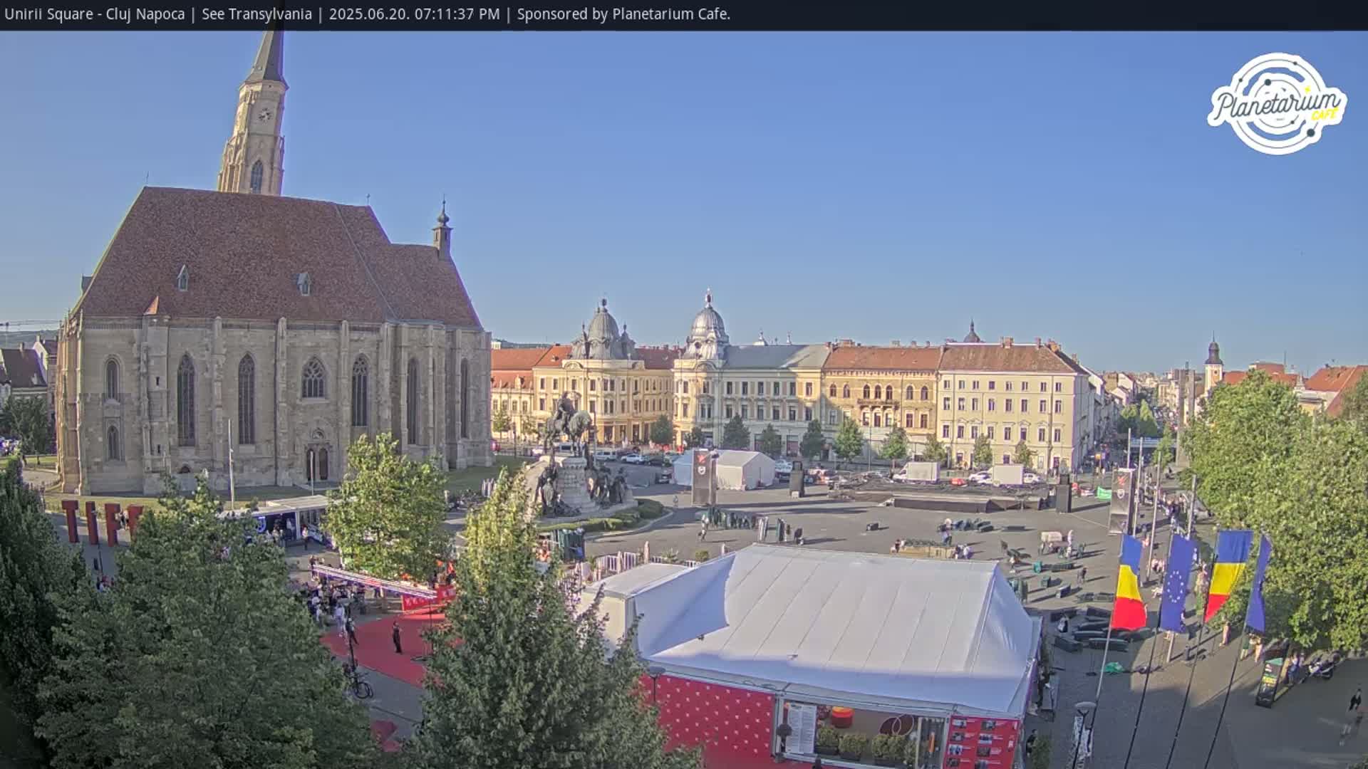 A large town square with a church, several buildings, a statue, and tents, under a clear blue sky.