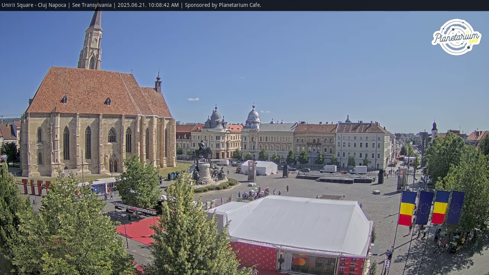 A large town square on a sunny day features a prominent church, several multi-story buildings, a statue, and several temporary structures.