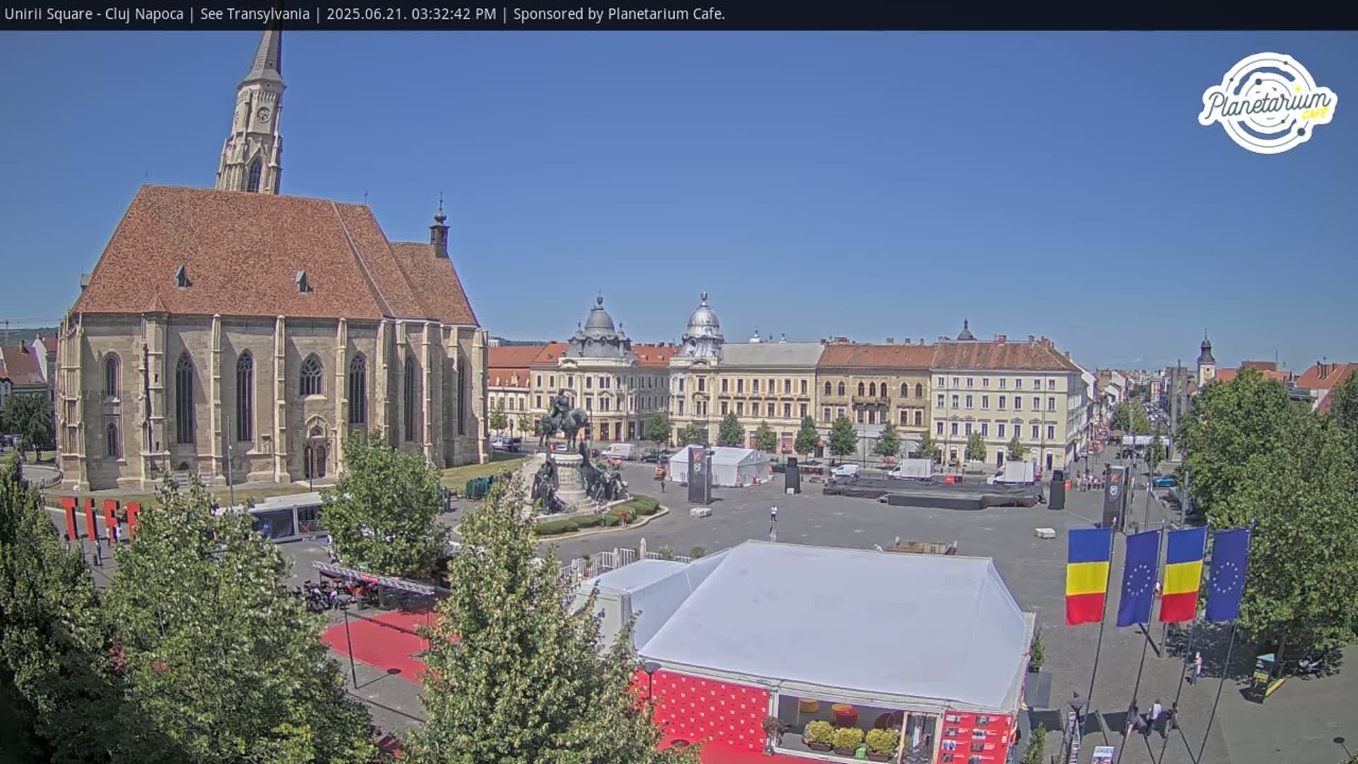 A large square with a church and several buildings is visible under a clear blue sky, with various tents and flags set up in the square.