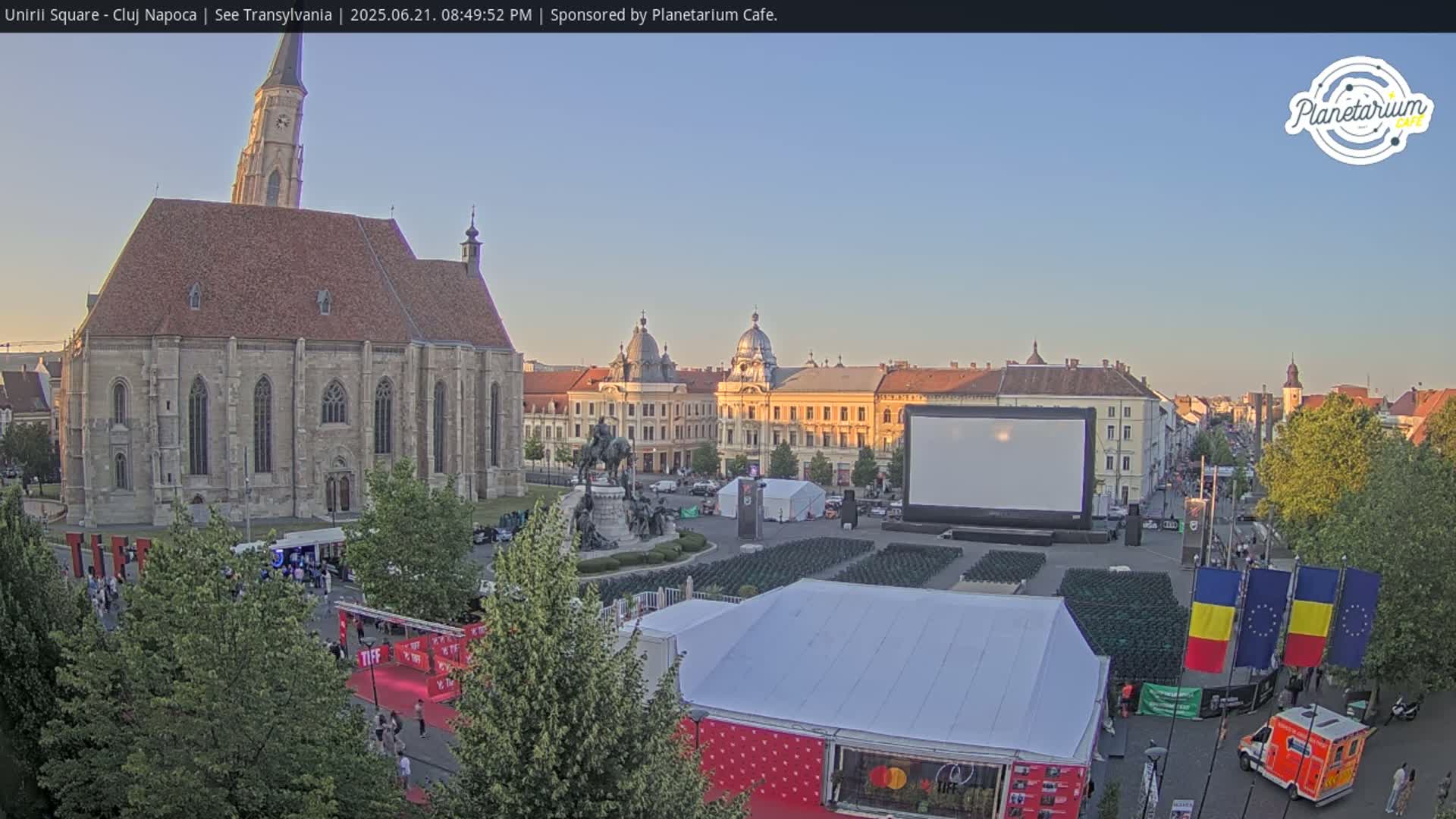 A large town square at sunset features a church, buildings, a large screen, and numerous chairs set up for an outdoor event.