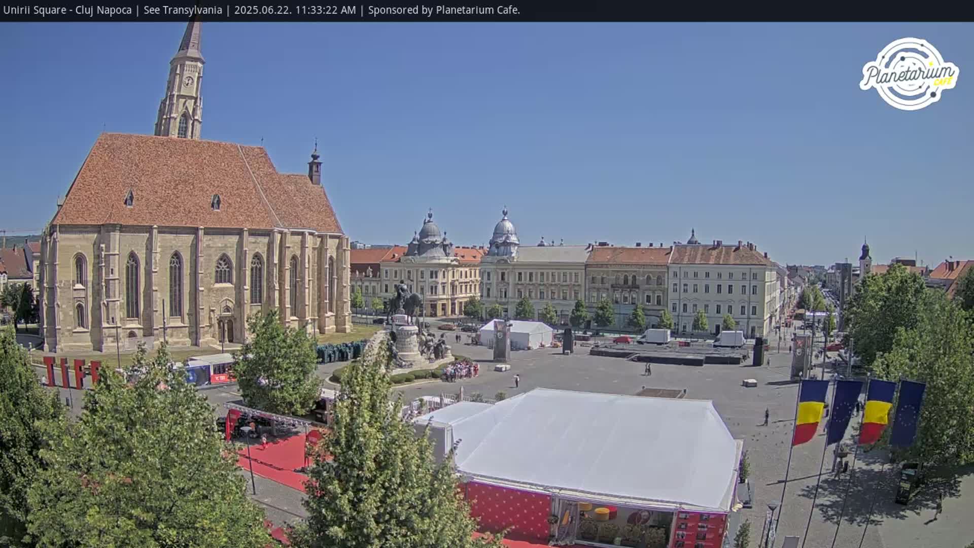 A large town square on a sunny day, featuring a large church, other buildings, a statue, and several tents.
