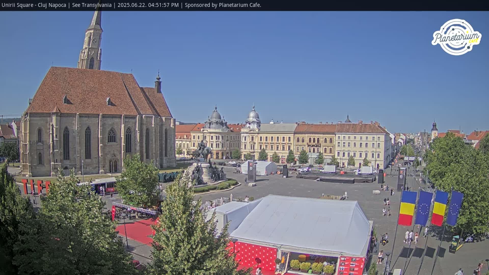 A large town square with a prominent church, several buildings, a statue, and temporary tents under a clear blue sky.