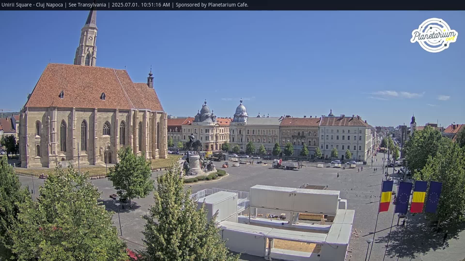 A large town square on a sunny day, featuring a prominent church, several other buildings, a statue, and some temporary structures in the center.