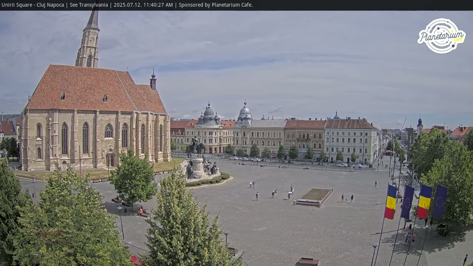 A large town square, under a partly cloudy sky, features a large church, a monument, and several multi-story buildings.