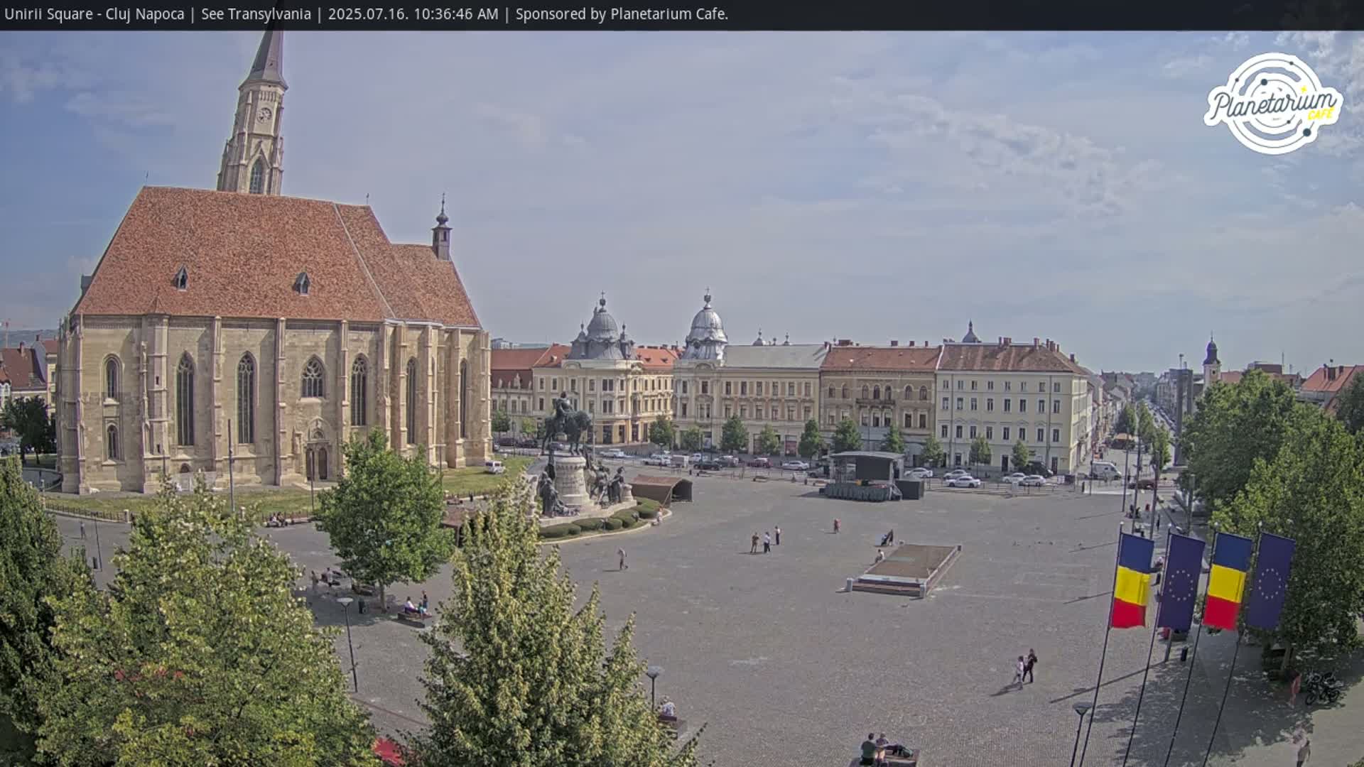 A large town square on a partly cloudy day, features a large church, several other buildings, a statue, and several Romanian and European Union flags.
