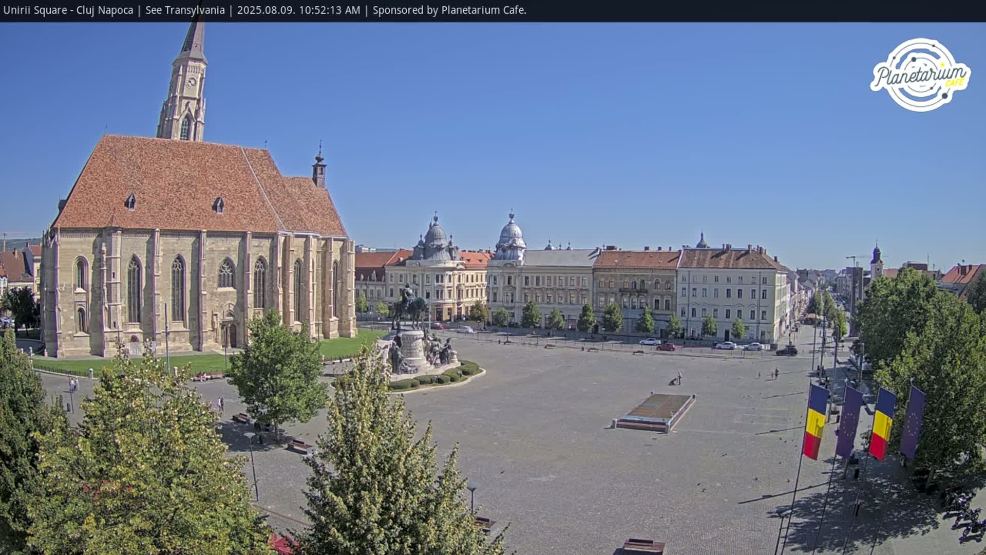 A large, mostly empty town square on a sunny day features a prominent church, a statue, and surrounding buildings, with Romanian flags waving in the foreground.