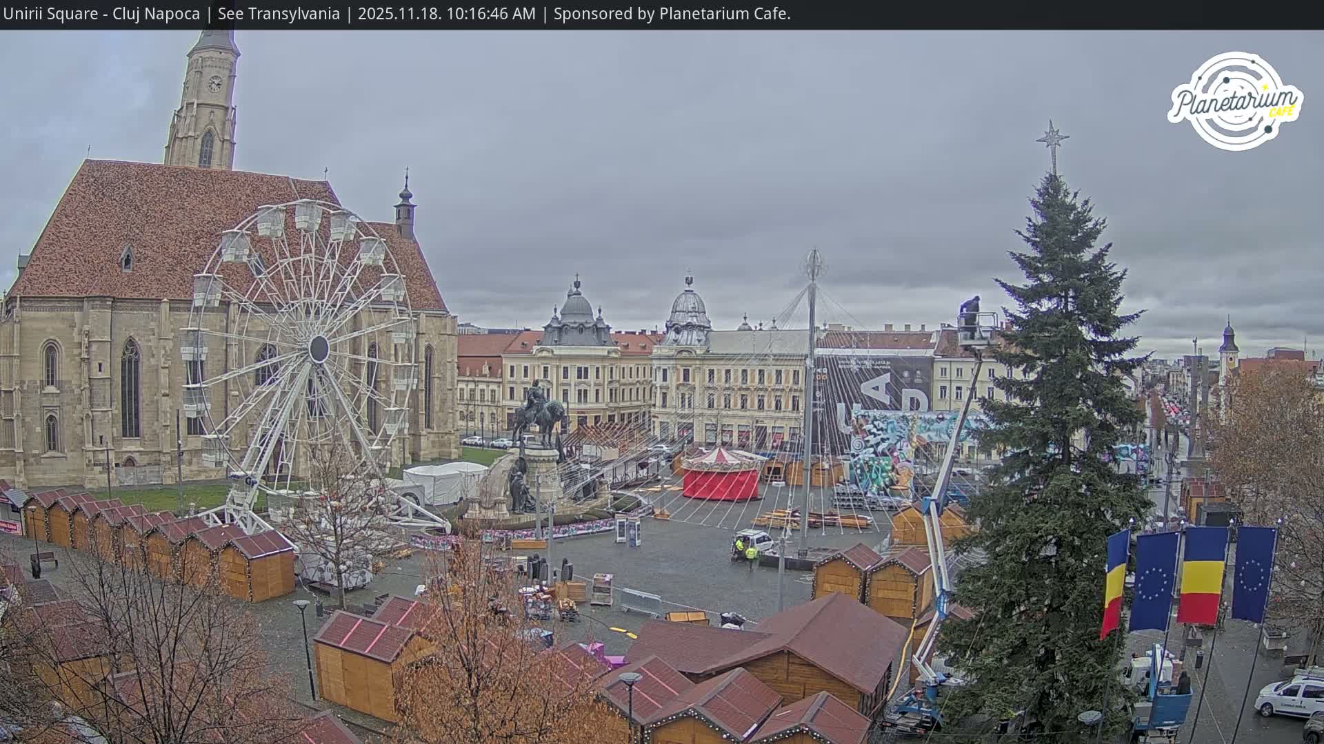 An overcast day in a European city square reveals a large Ferris wheel next to a historic church, an equestrian statue, numerous wooden market stalls, and a Christmas tree being decorated by a worker on a lift.