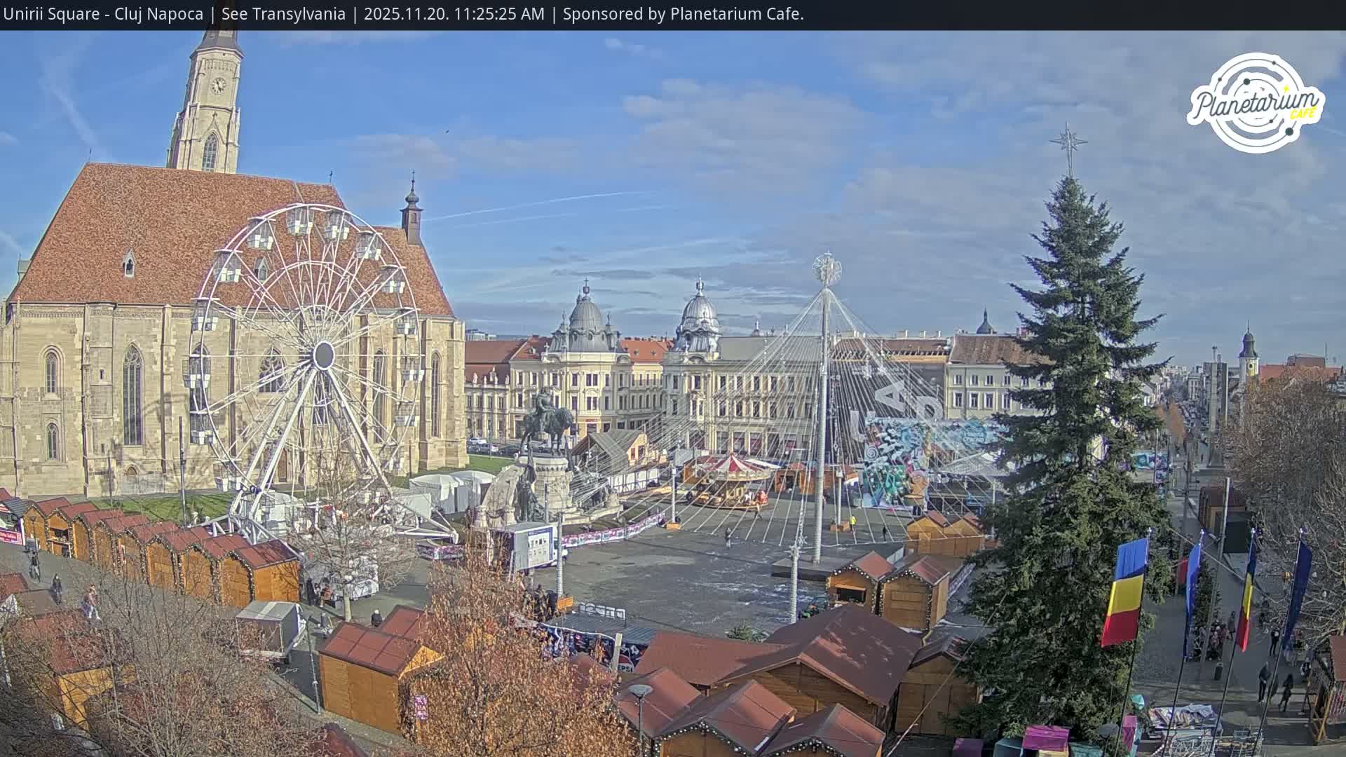 A festive outdoor market, featuring a large Ferris wheel, a tall Christmas tree, and numerous stalls, is set up in a historic city square under a clear, sunny sky, with a grand church visible on the left and stately buildings in the background.