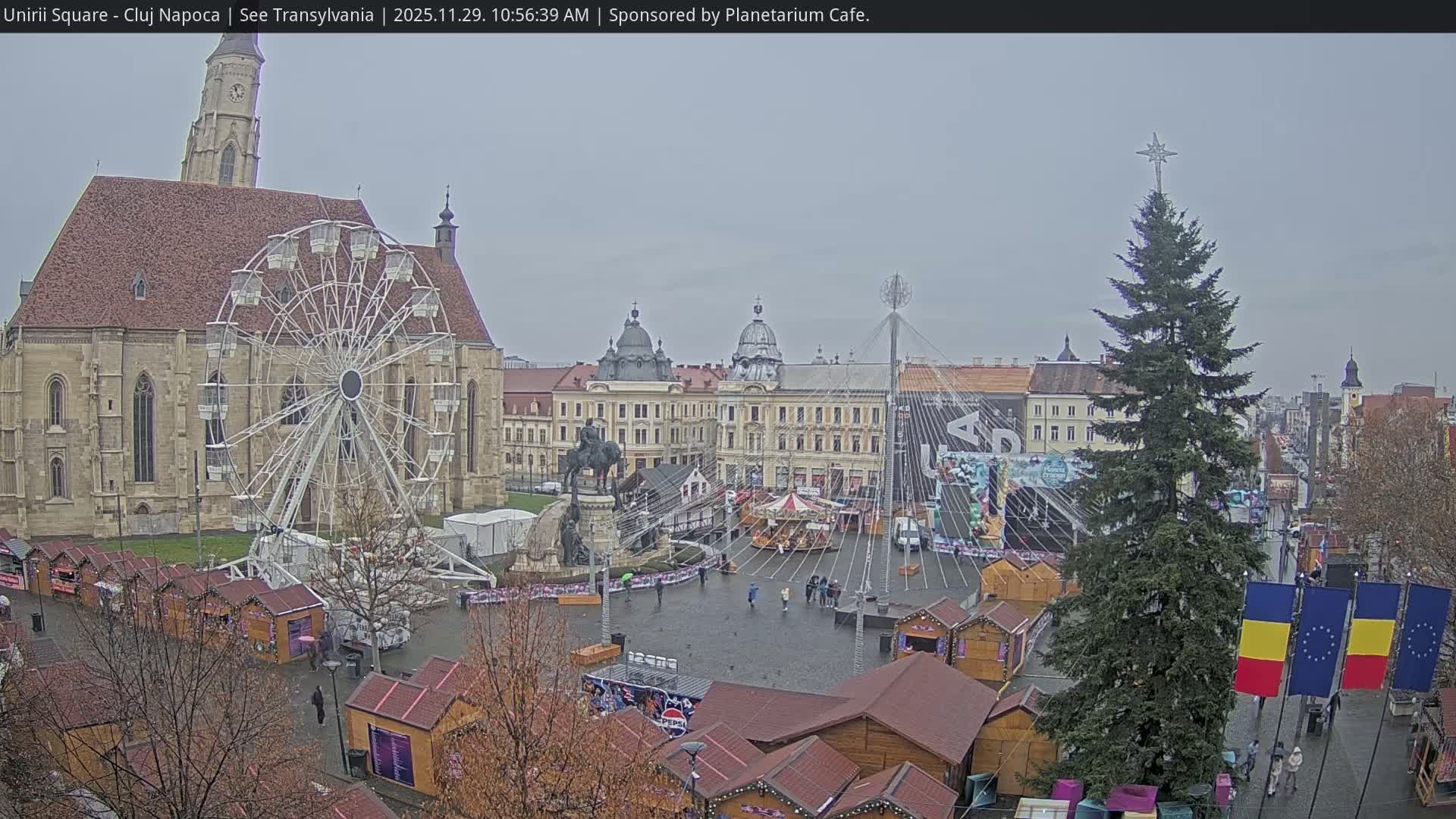 An overcast sky hangs over a large city square, filled with a Ferris wheel, a tall Christmas tree topped with a star, numerous market stalls, and a carousel, flanked by a grand church and other historic buildings, with an equestrian statue at its center and a few pedestrians.