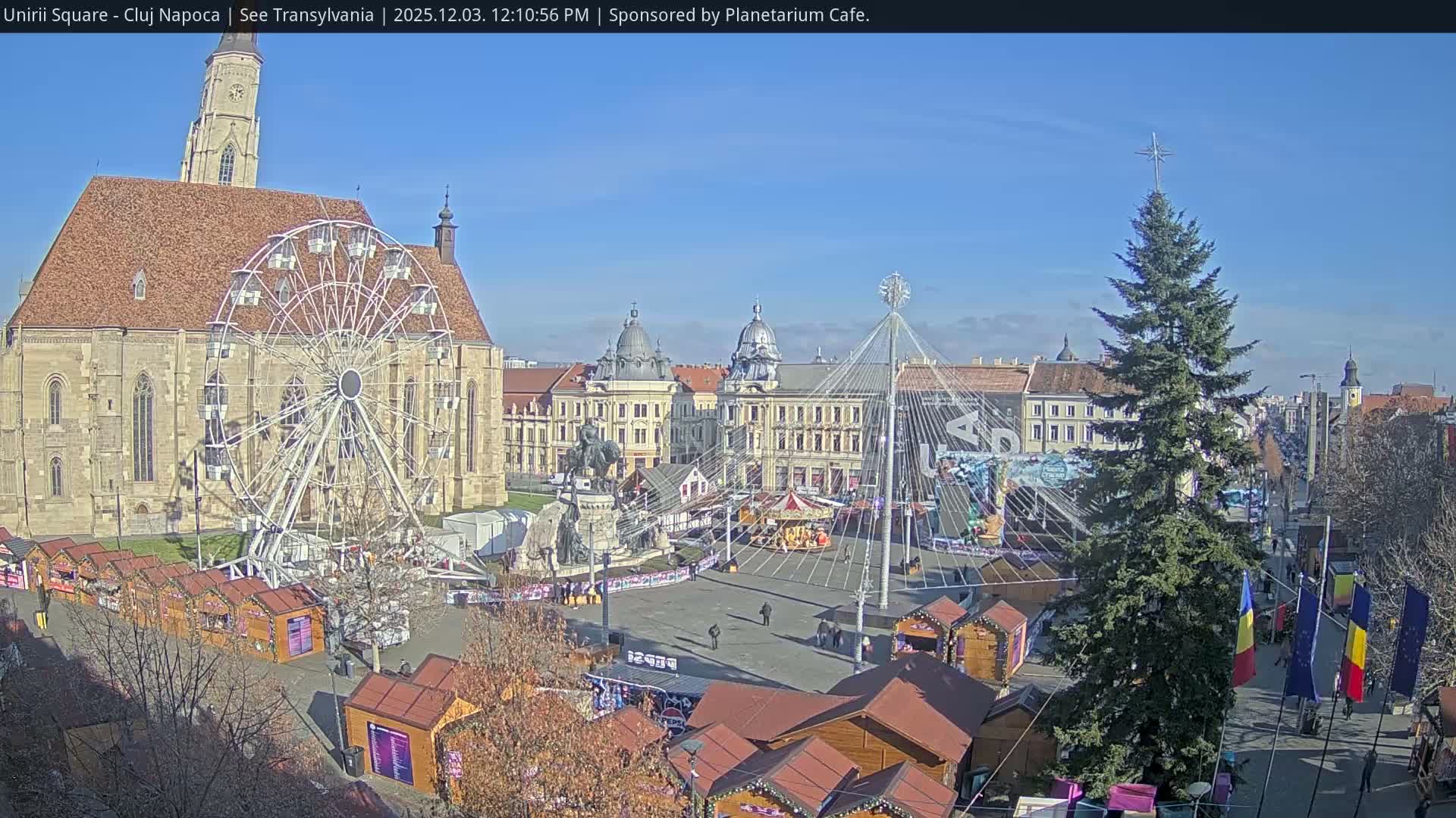 A large city square configured as a Christmas market is bathed in bright sunlight under a clear blue sky, featuring a grand Gothic church, a towering Ferris wheel, numerous wooden stalls, an equestrian statue, and a large decorated Christmas tree.