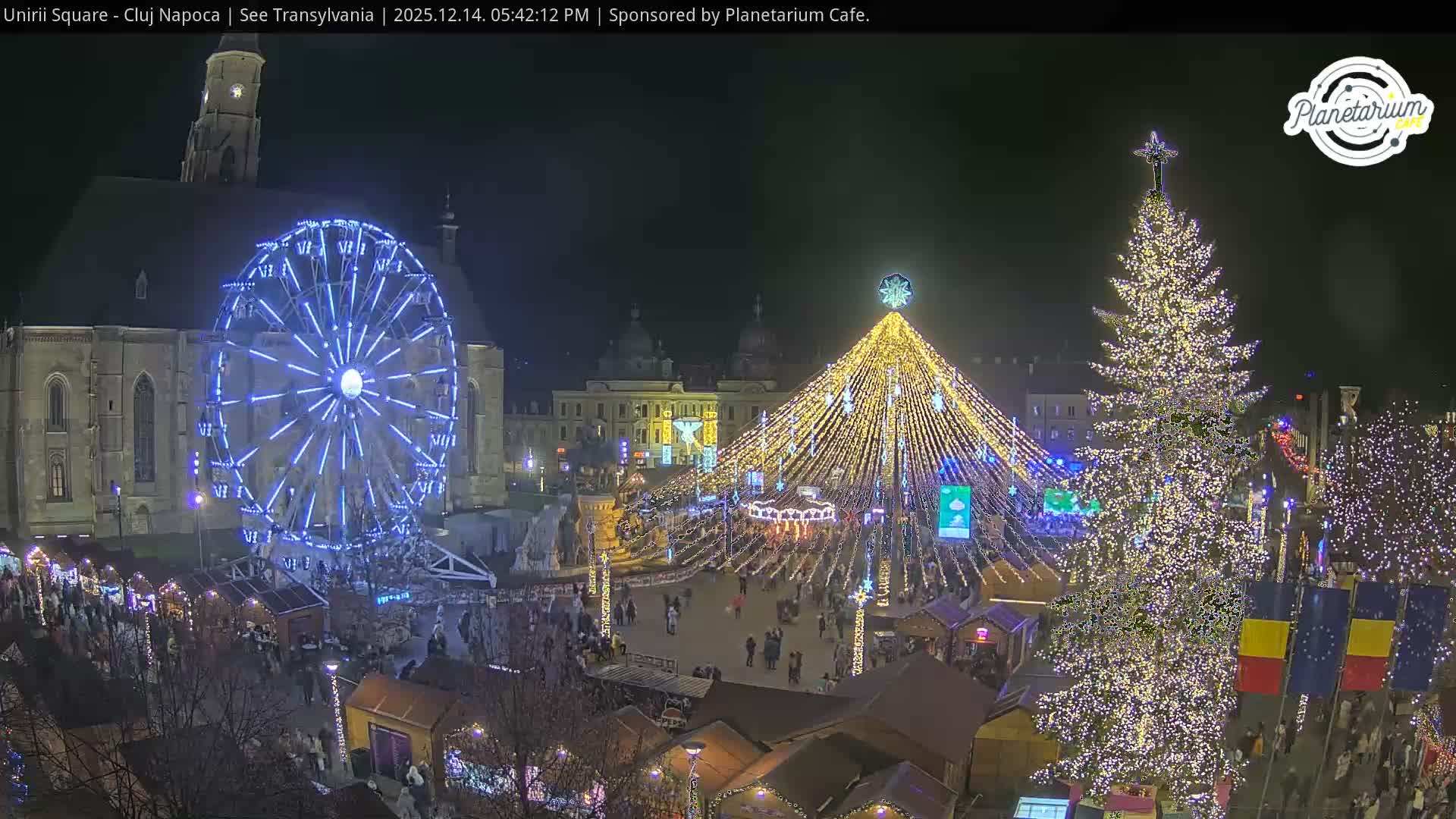A large city square configured as a Christmas market is bathed in bright sunlight under a clear blue sky, featuring a grand Gothic church, a towering Ferris wheel, numerous wooden stalls, an equestrian statue, and a large decorated Christmas tree.