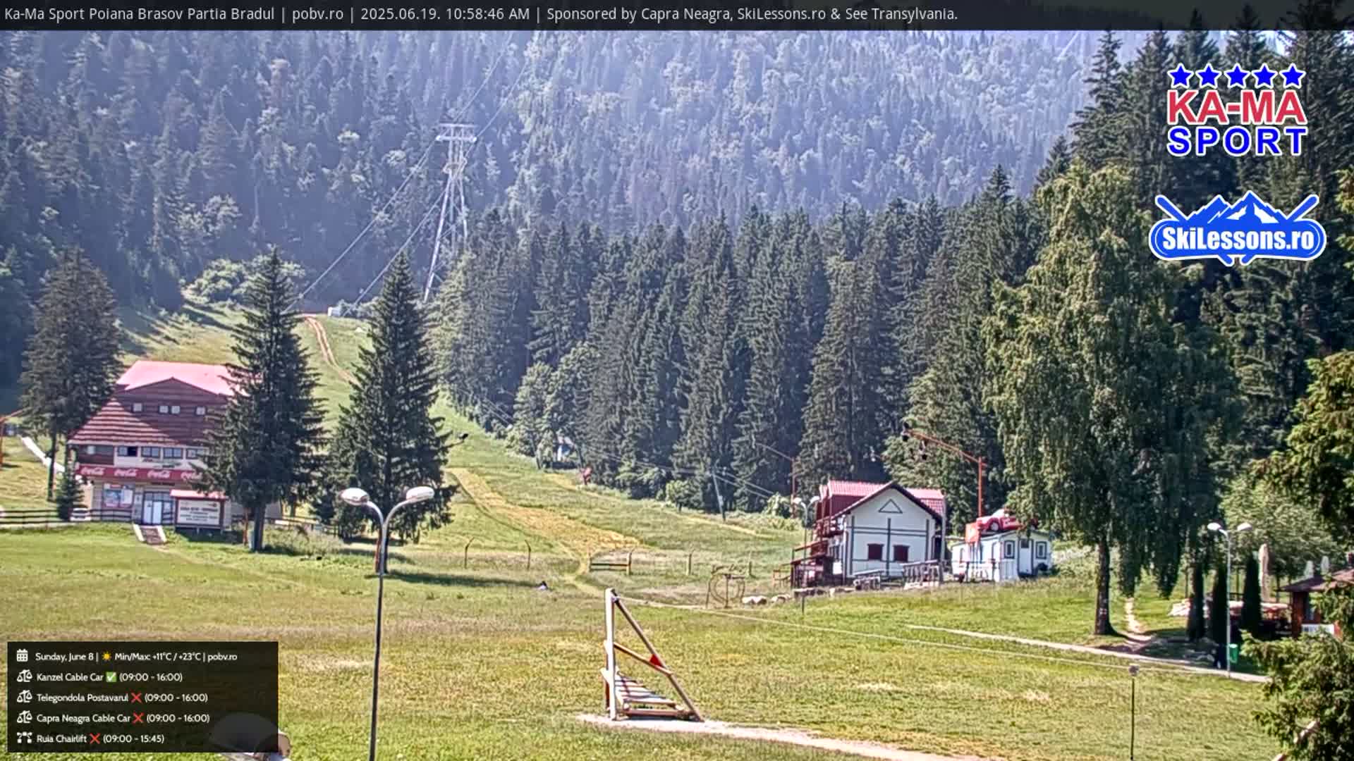 A sunny day at a ski resort shows green hills, evergreen trees, and several buildings, with a chairlift visible in the distance.