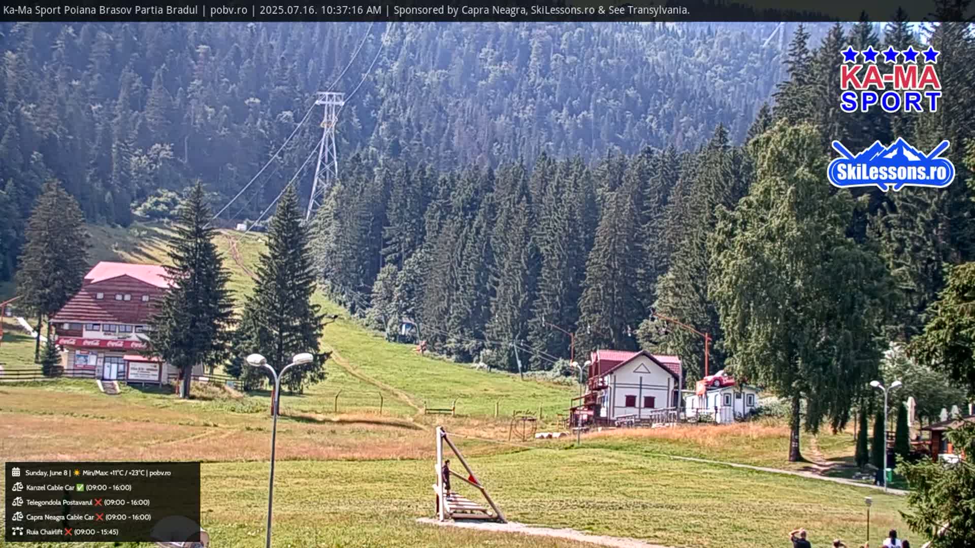 A sunny day at a mountain resort shows a chairlift, buildings, and grassy hillsides surrounded by a dense evergreen forest.