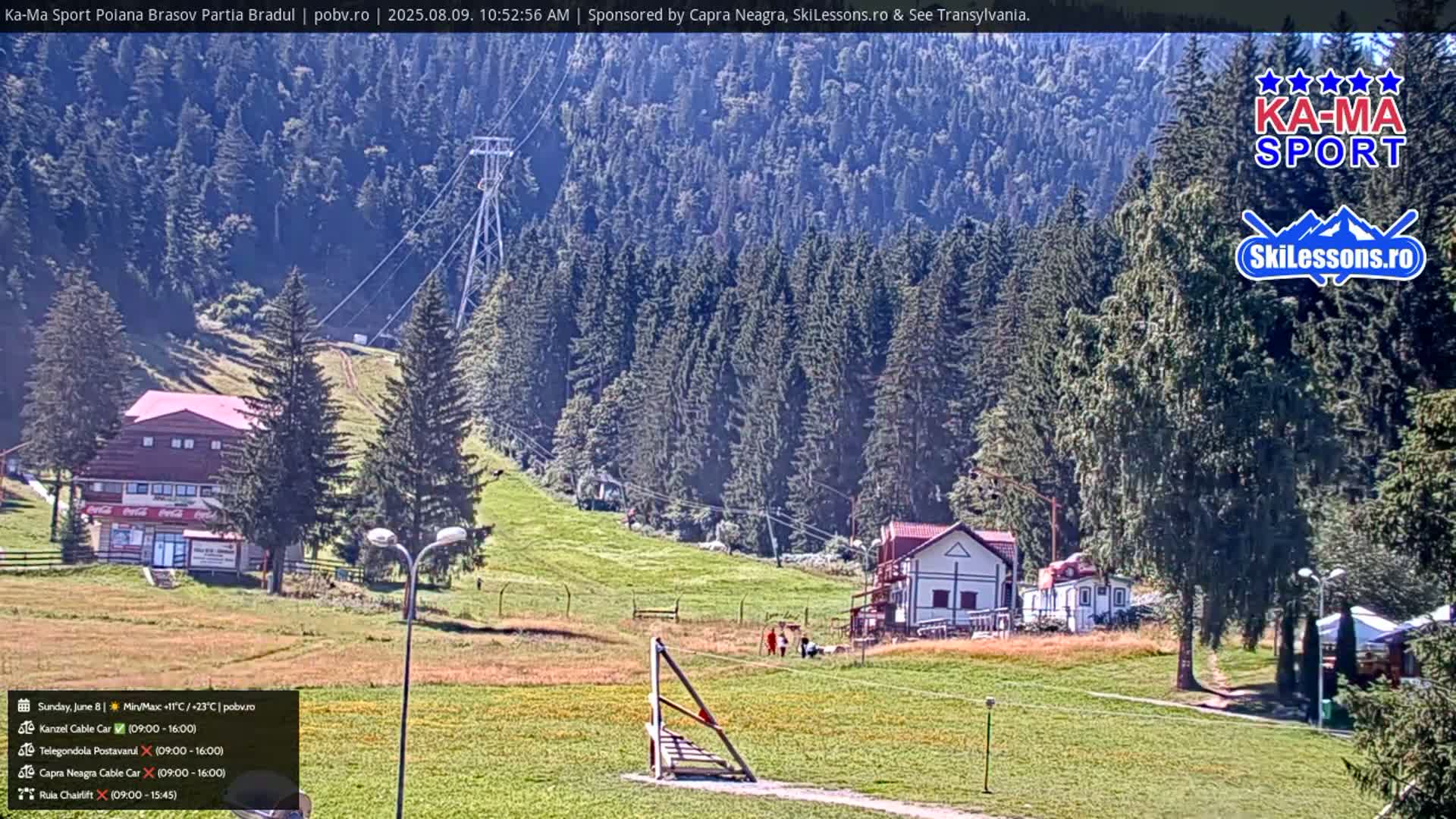A sunny day in a mountainous area shows a grassy field with a few buildings, a ski lift, and a forest in the background.
