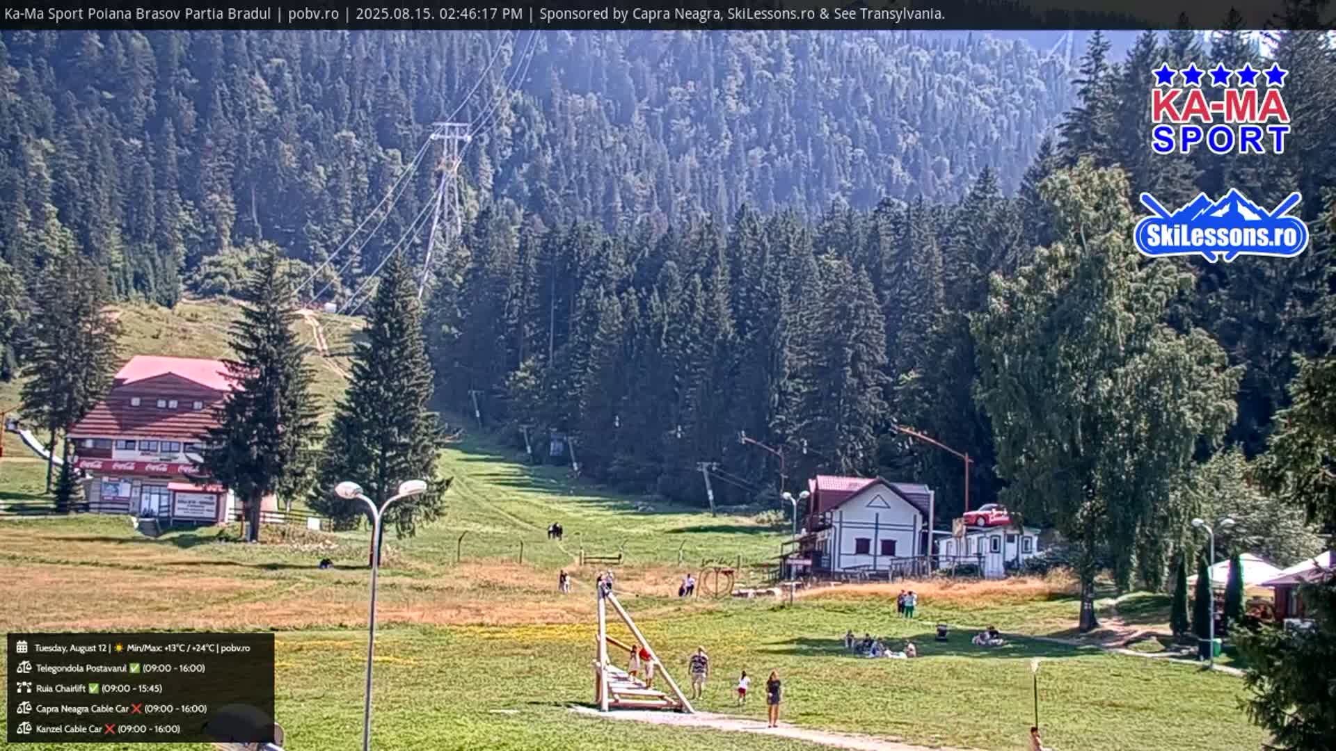 A sunny, outdoor view of a grassy area with buildings, a chairlift, and a forest in the background.
