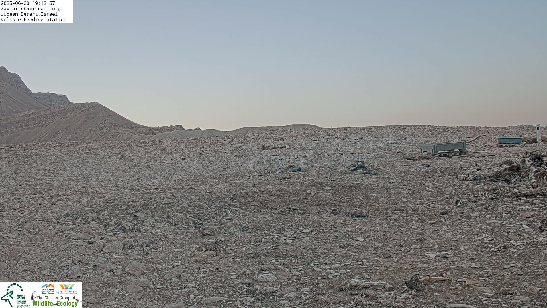 A rocky, arid landscape under a pale sky features two dark gray containers and scattered debris near a low hill.