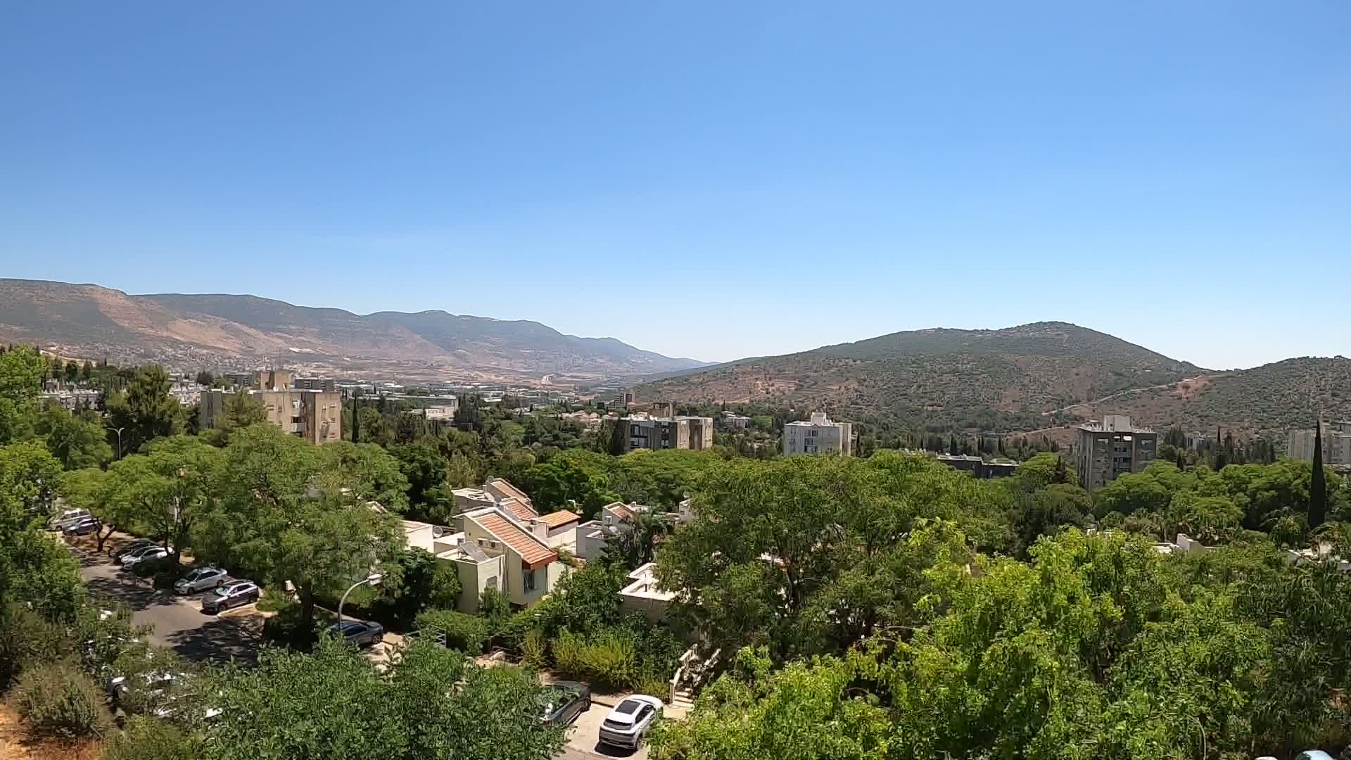 A residential area nestled among lush green trees and shrubs overlooks a valley with distant mountains under a clear blue sky.