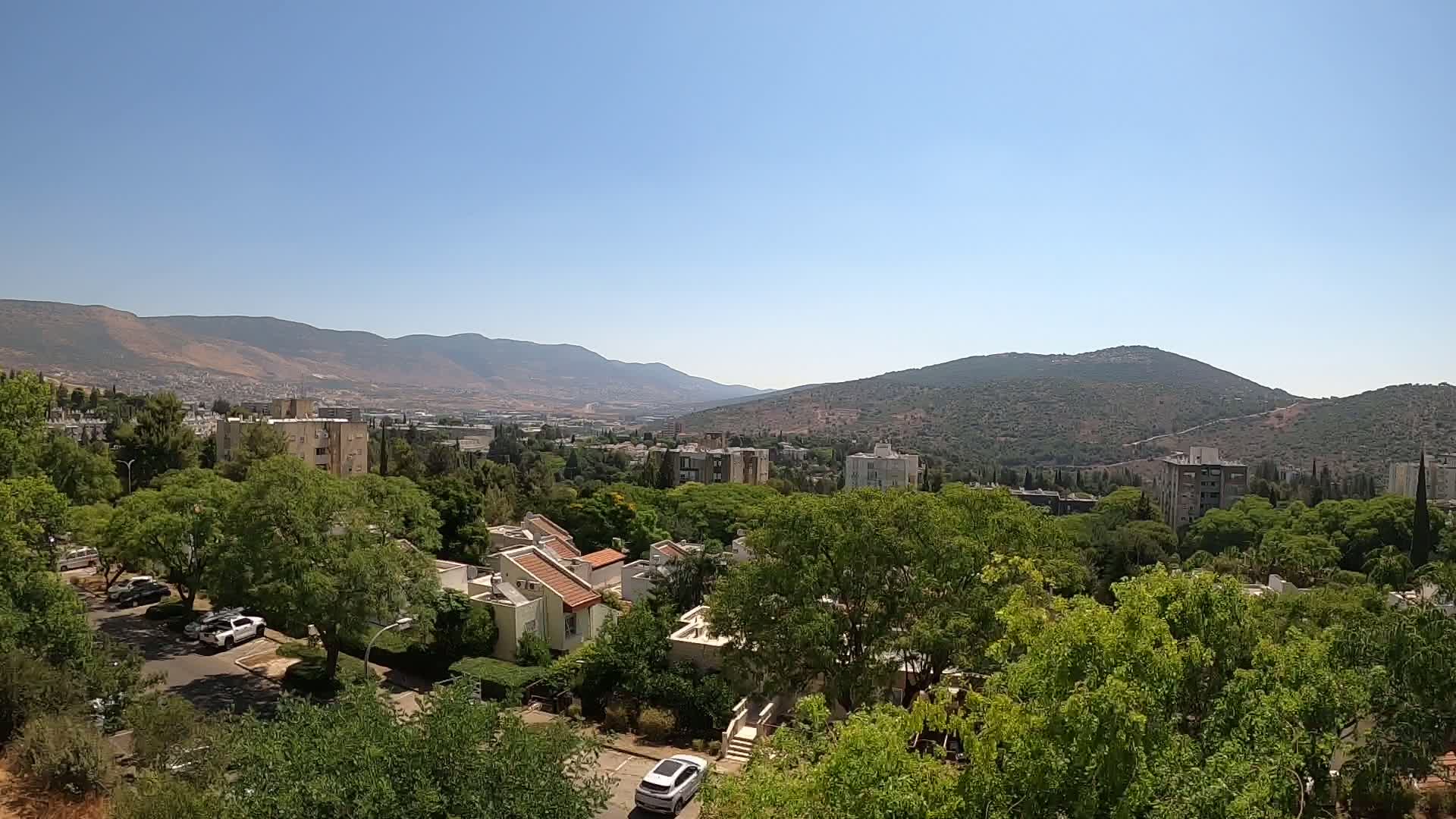 A residential area nestled among lush green trees and shrubs, overlooks a valley with distant mountains under a clear, sunny sky.