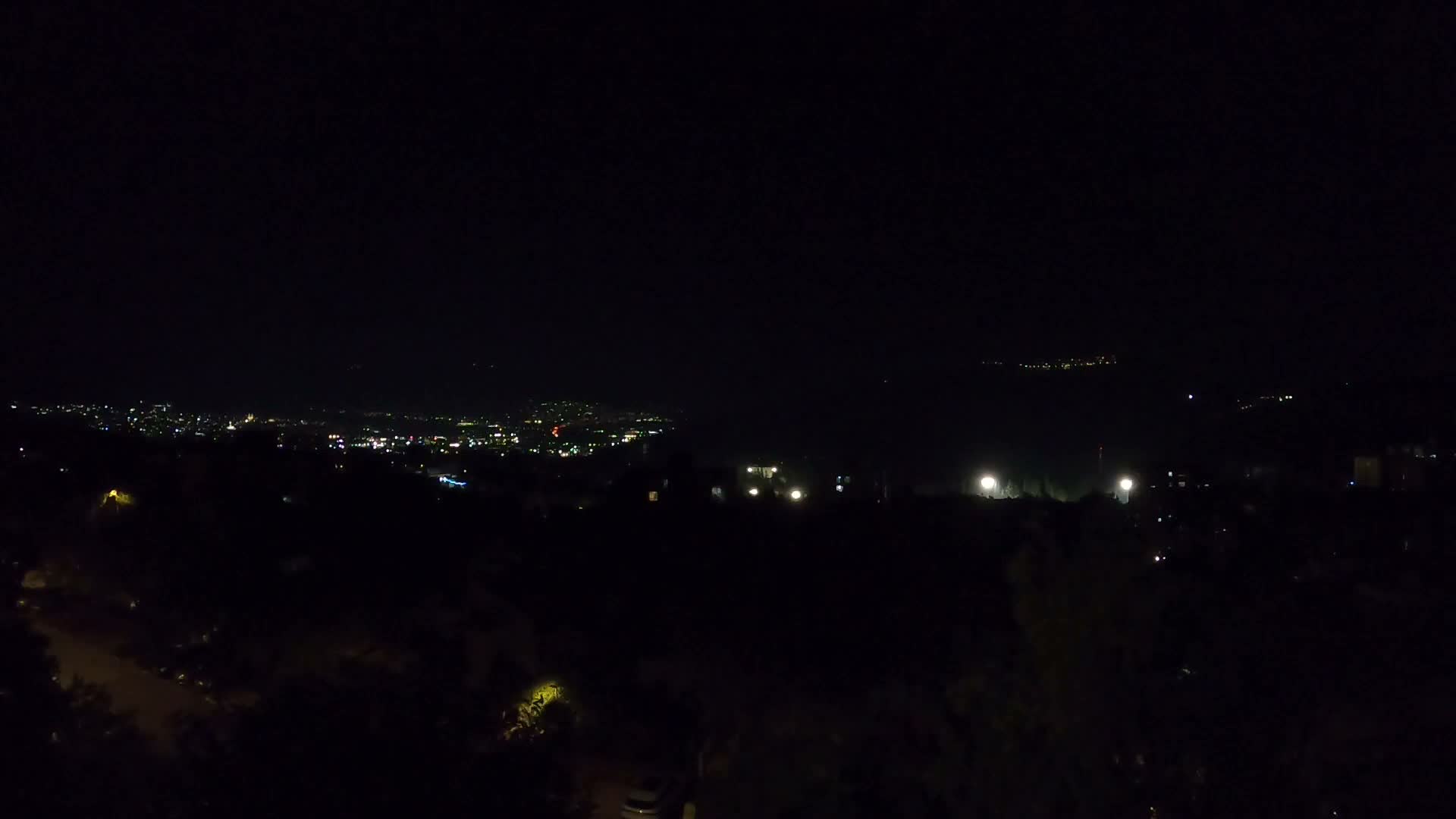 A residential area nestled among lush green trees and shrubs, overlooks a valley with distant mountains under a clear, sunny sky.