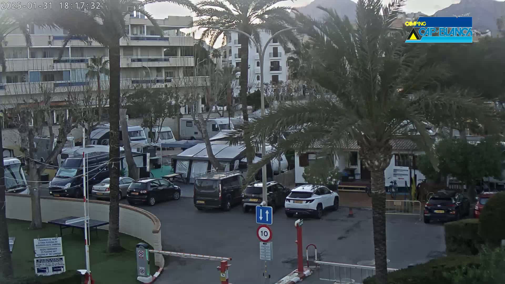 A parking lot filled with cars and campervans is situated near a multi-story building under an overcast sky.