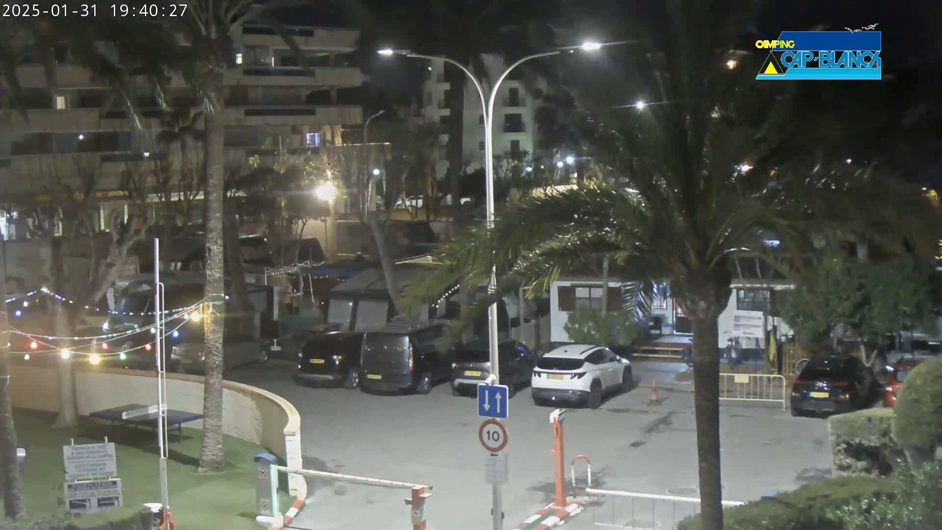 A nighttime view of a parking lot with several cars, palm trees, and buildings under a clear sky.