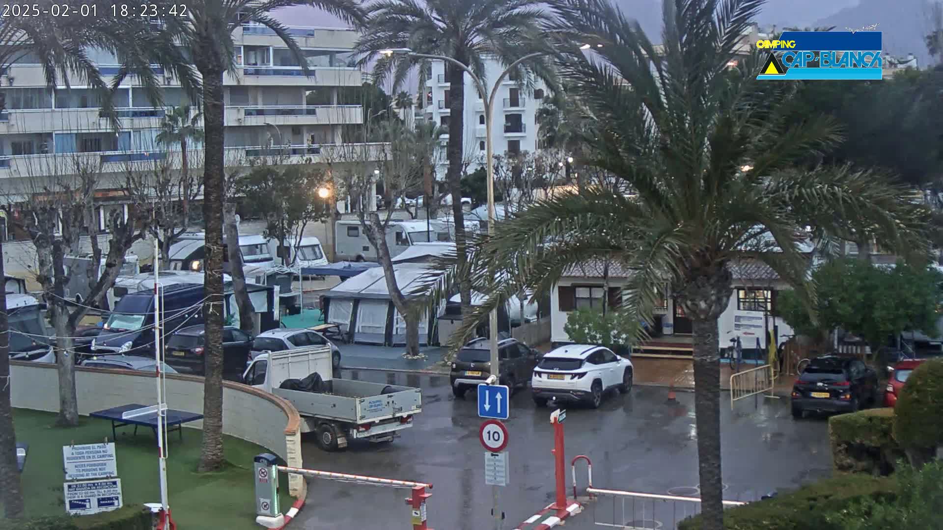 A wet campground with numerous RVs, cars, and tents is situated near apartment buildings under a cloudy sky.
