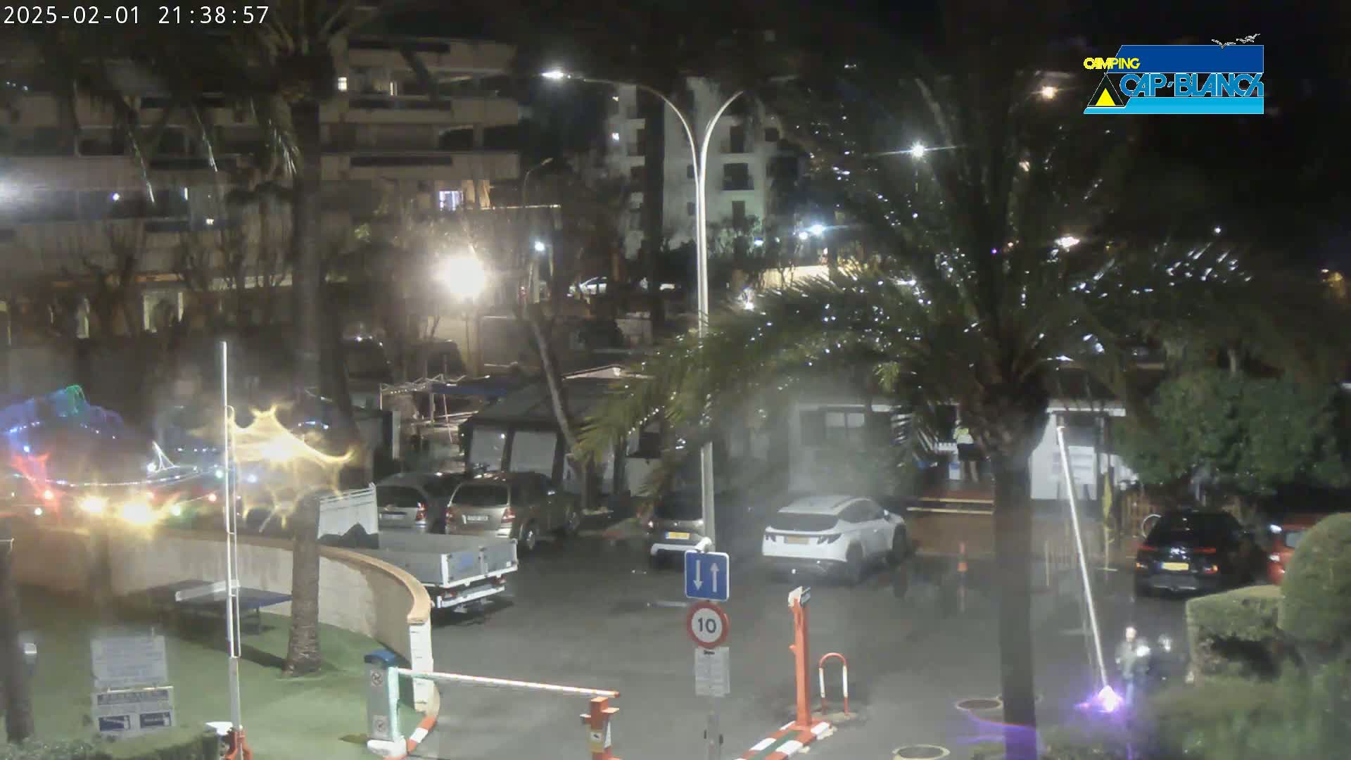 A nighttime scene shows a parking lot with cars and RVs near buildings, palm trees, and streetlights under wet conditions.