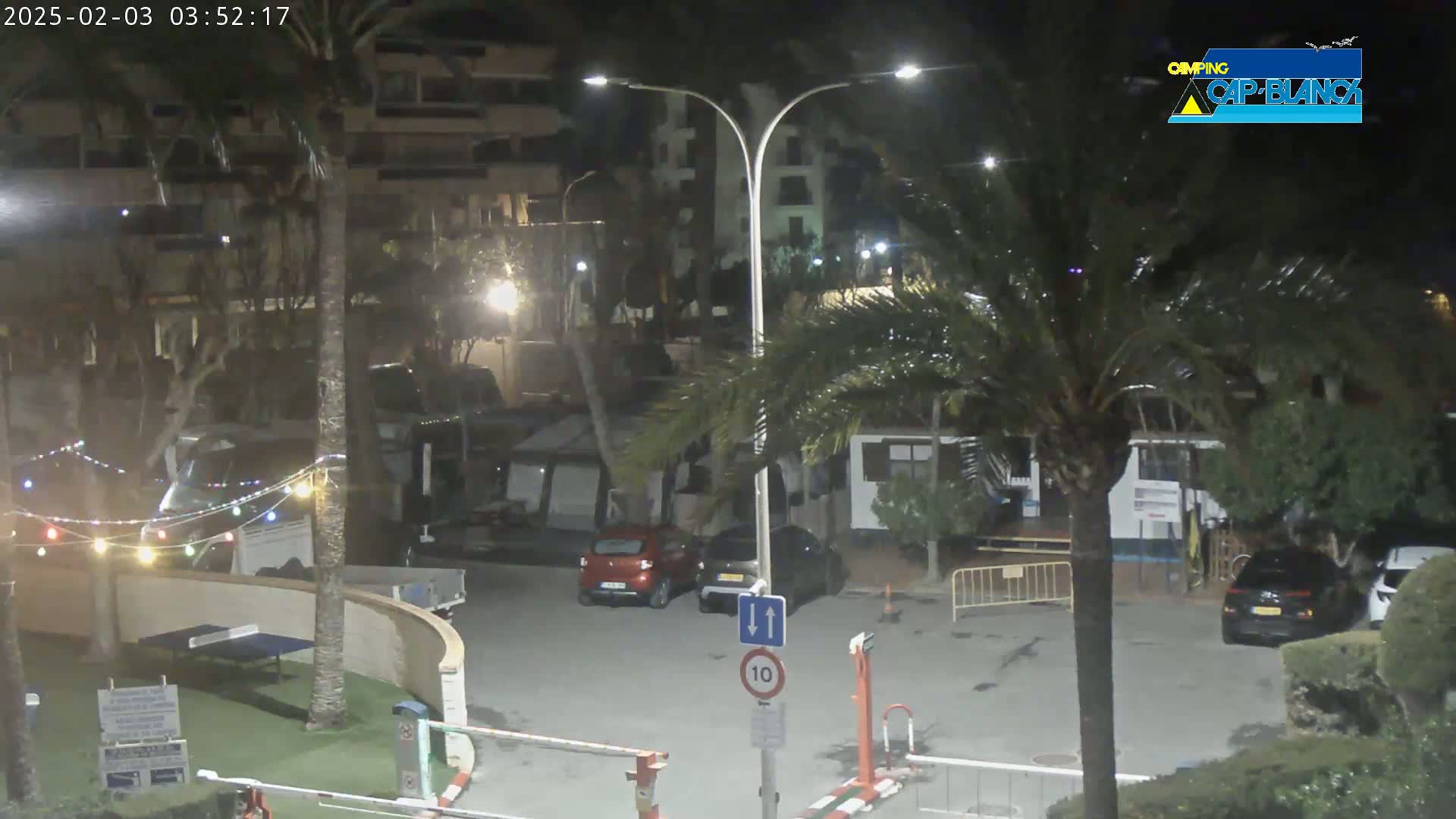 A nighttime view of a parking lot with several cars, palm trees, and buildings illuminated by streetlights.
