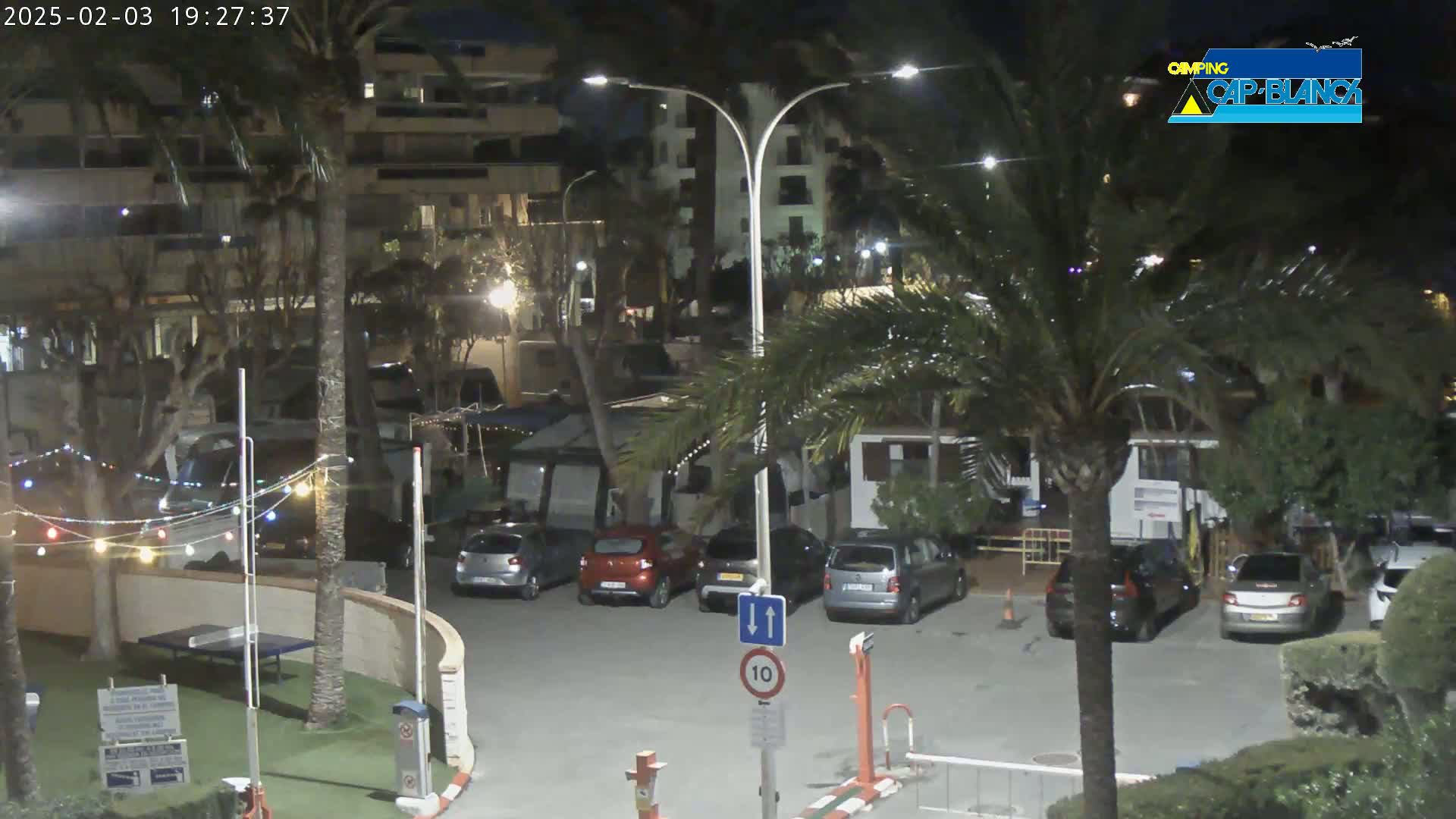 A nighttime view of a parking lot with several cars parked, surrounded by palm trees, buildings, and string lights.