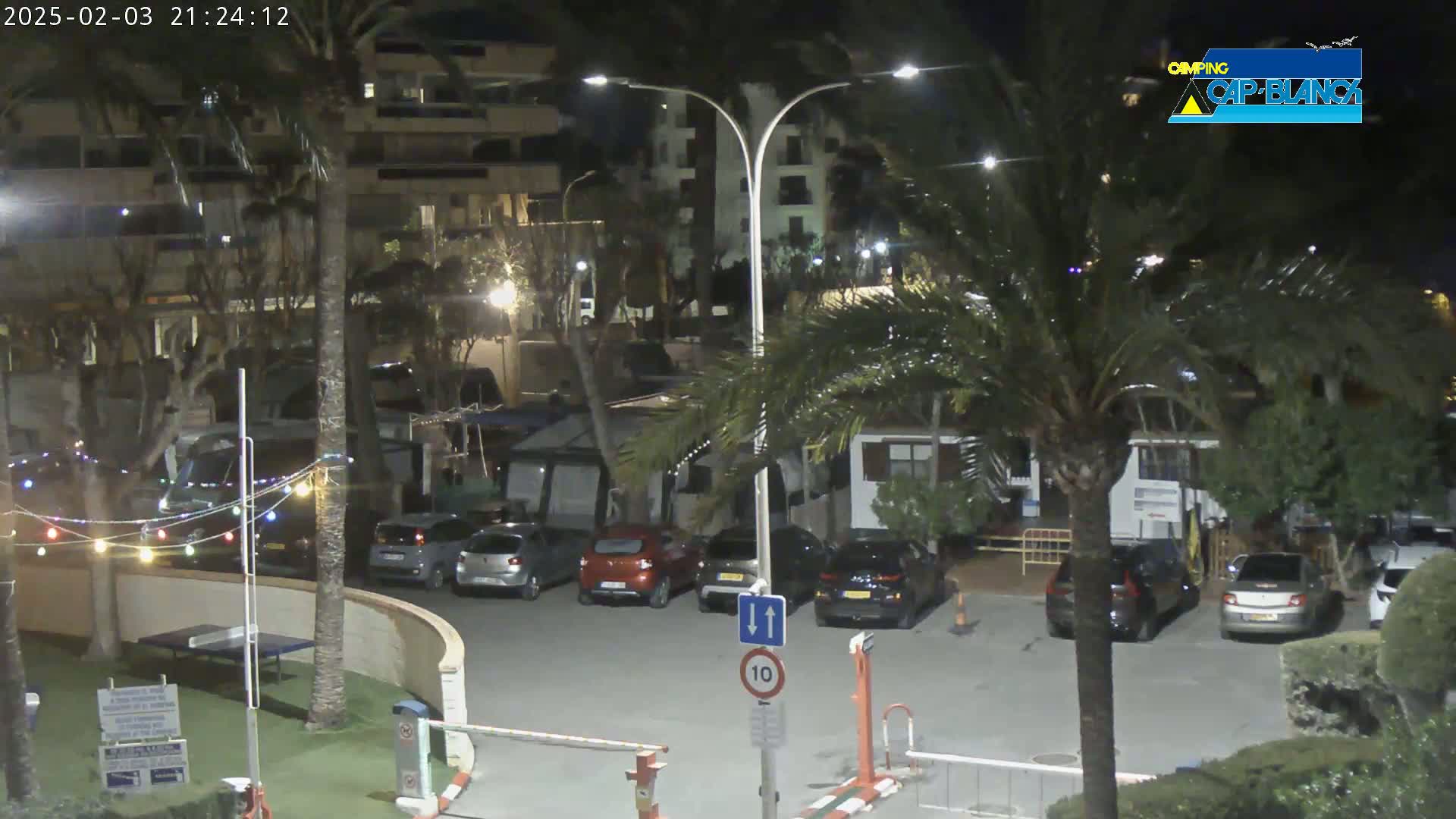 A nighttime view of a parking lot in a campground, surrounded by palm trees and buildings, with several cars parked and string lights illuminating the area.