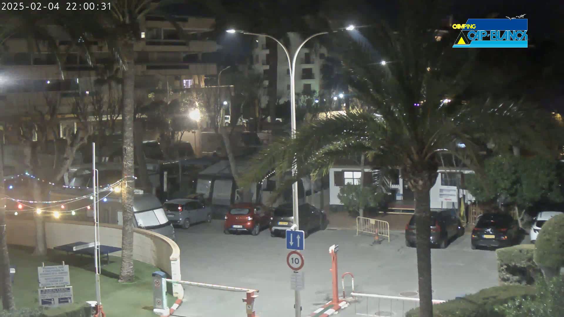 A nighttime view of a parking lot with cars and RVs, surrounded by buildings, palm trees, and string lights.