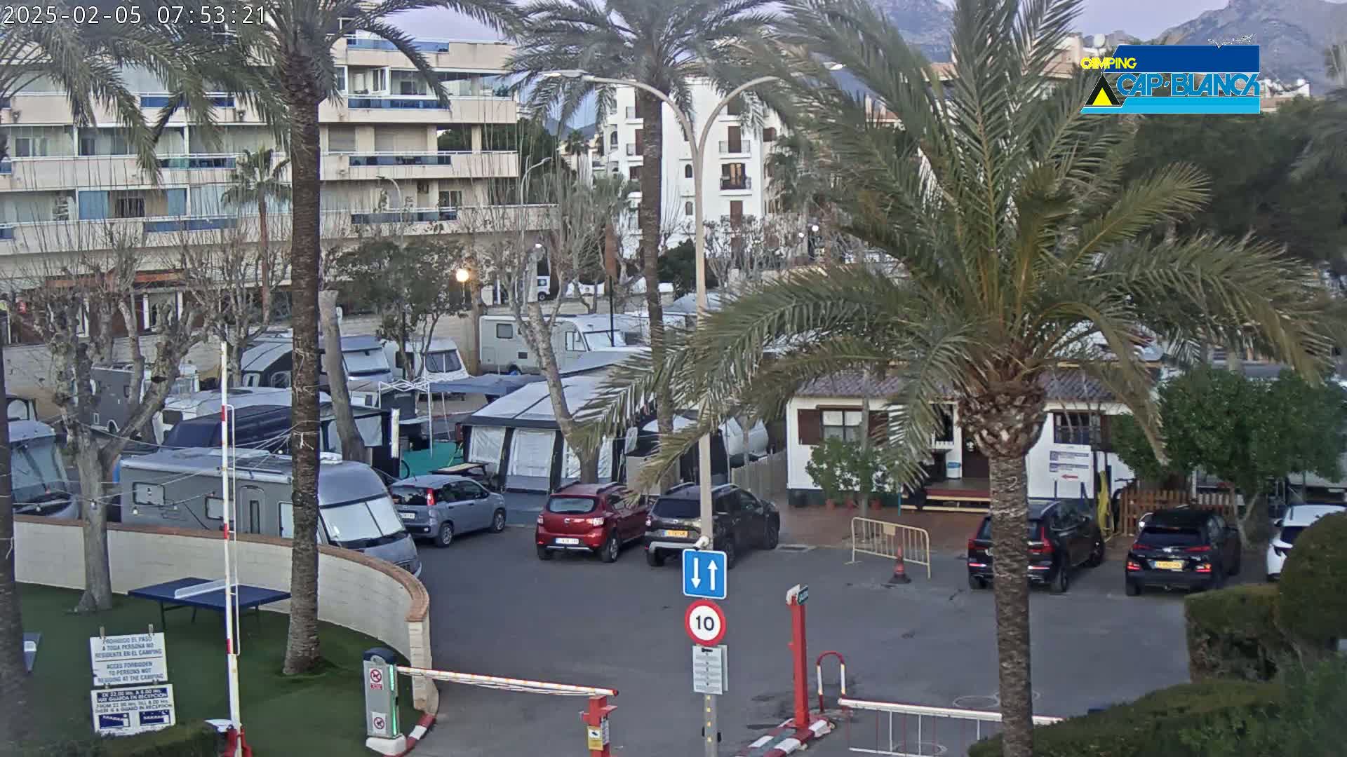 A parking lot filled with campervans and cars is situated near apartment buildings under a mostly cloudy sky, with palm trees and mountains visible in the background.