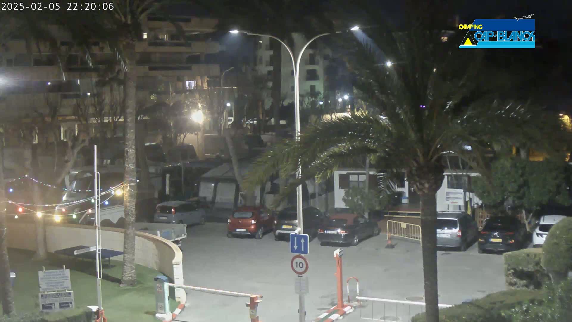 At night, under clear skies, several cars are parked in a lot near buildings and palm trees, with string lights visible in the foreground.