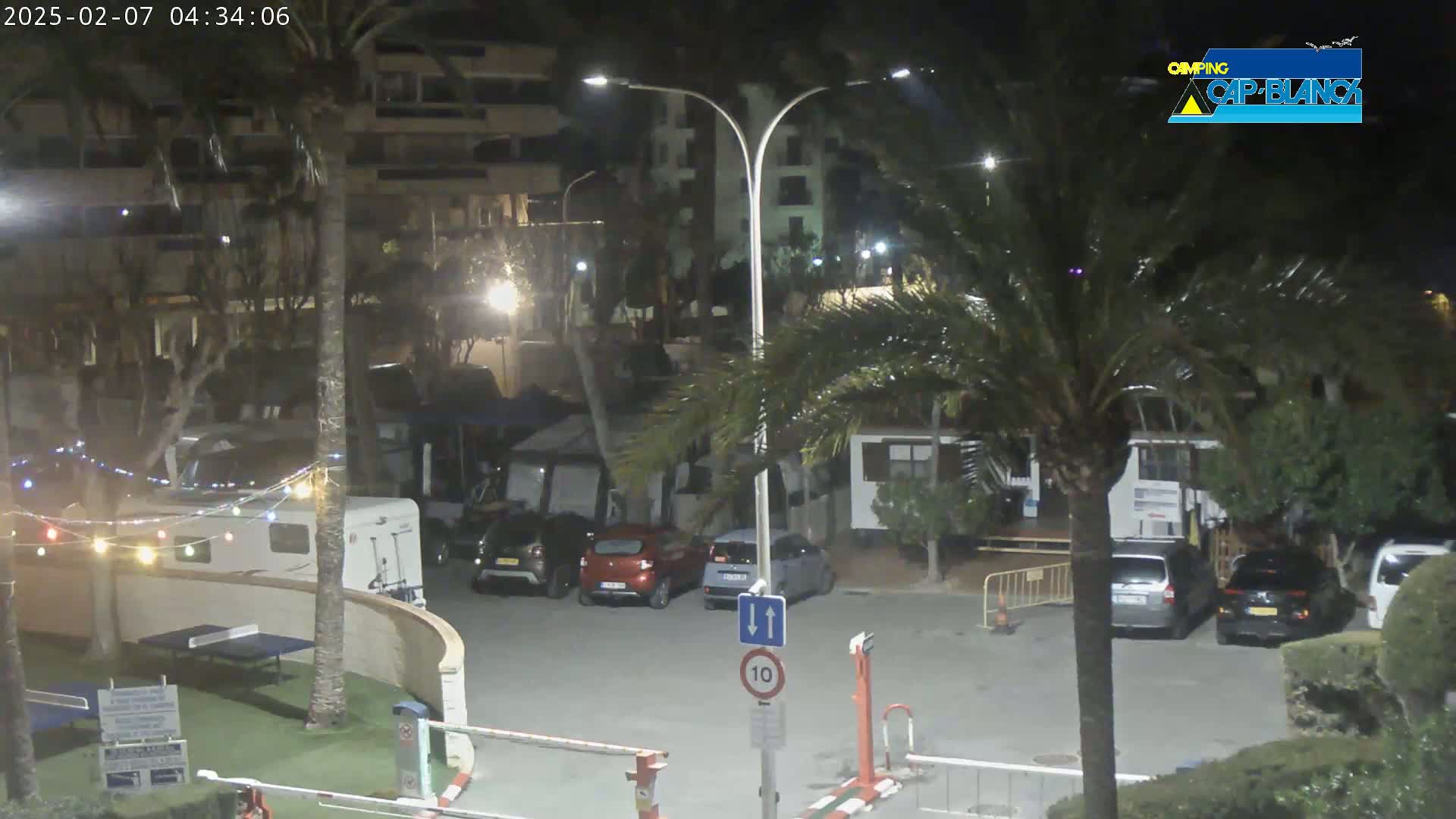 A nighttime view of a parking lot with several cars and RVs, string lights, and palm trees, under mostly clear skies.