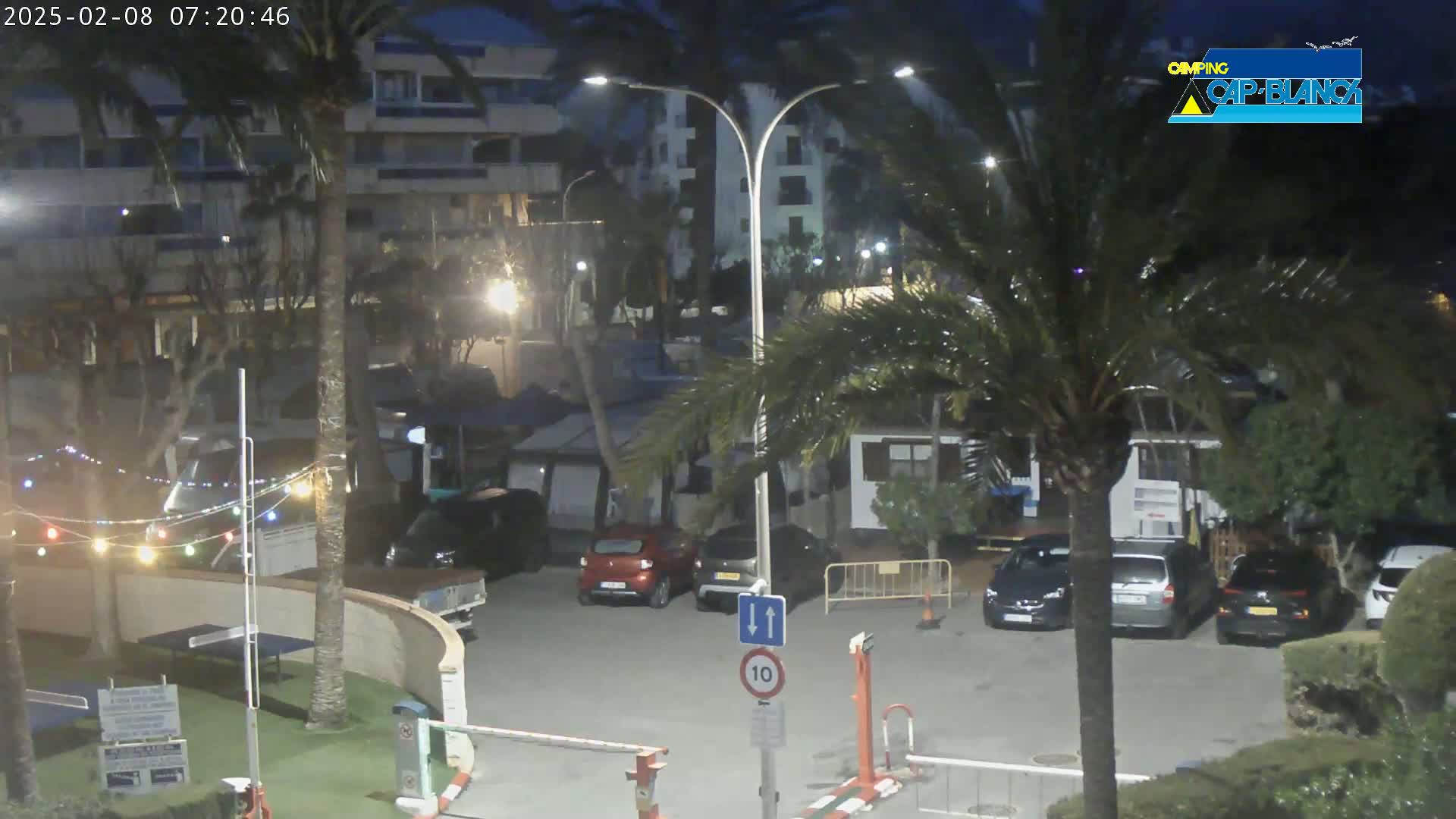 A nighttime parking lot scene with several cars parked, palm trees, and a building in the background, under partly cloudy skies.