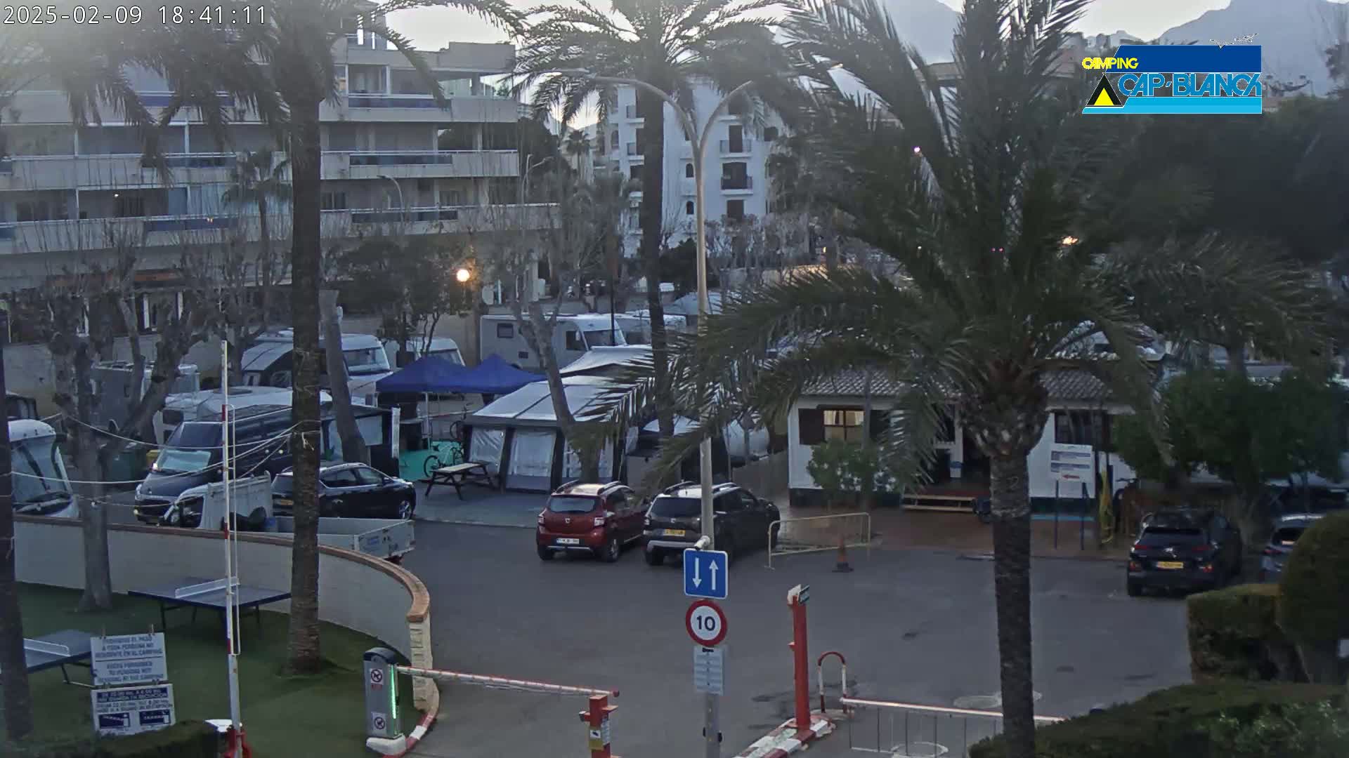 A parking lot with numerous cars, RVs, and tents is situated near a multi-story building under a hazy, overcast sky.