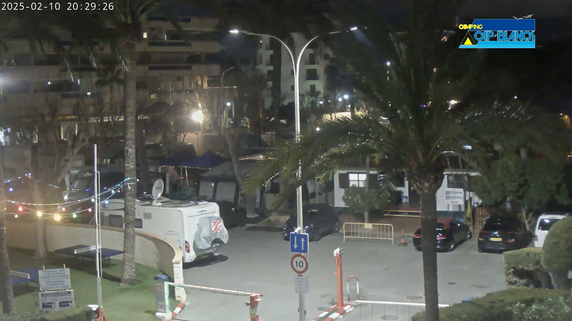 At night, under partly cloudy skies, a parking lot with several cars and recreational vehicles is illuminated by streetlights amidst palm trees and buildings.