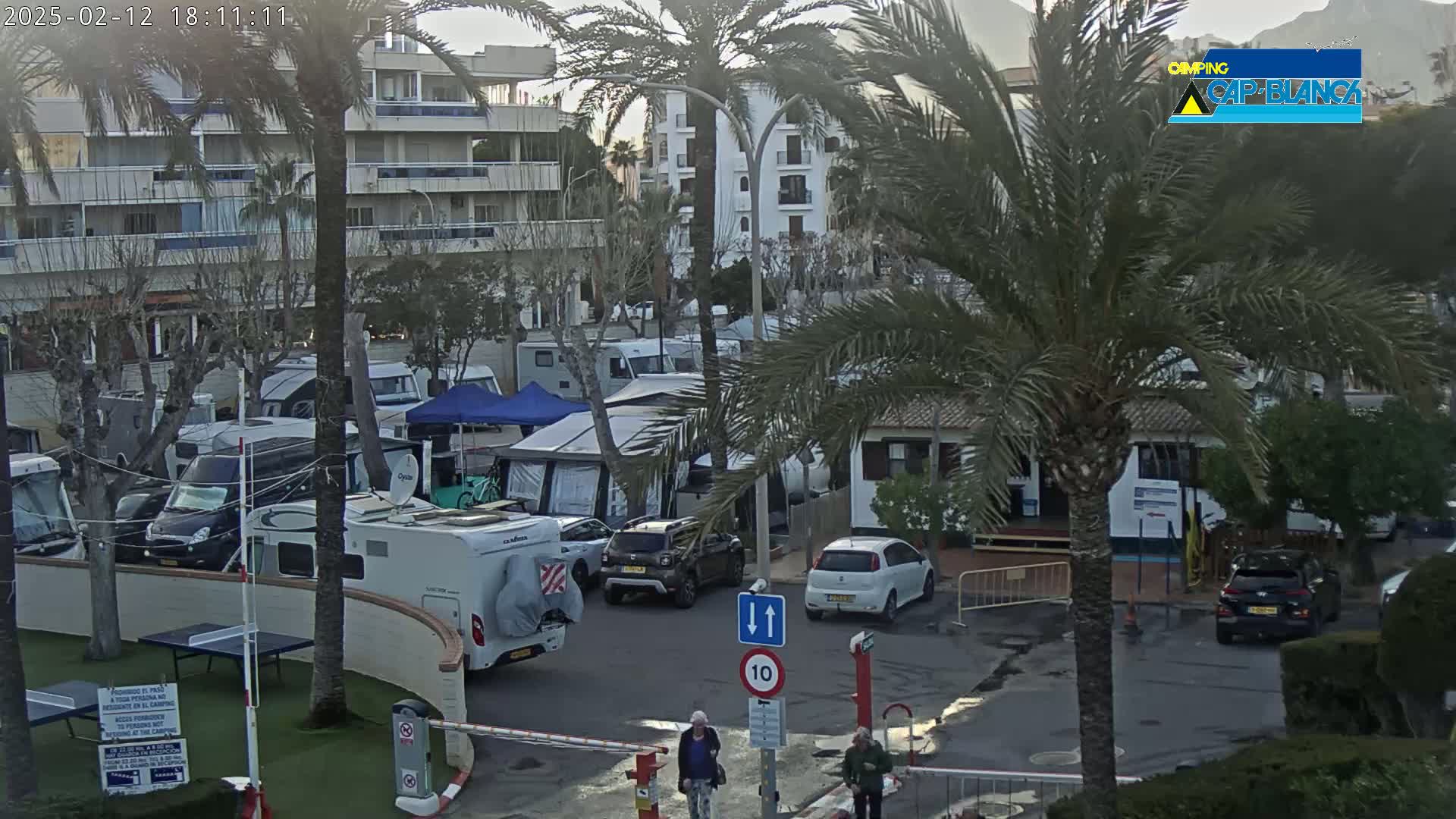 Numerous RVs and cars are parked in a campground next to apartment buildings under an overcast sky.